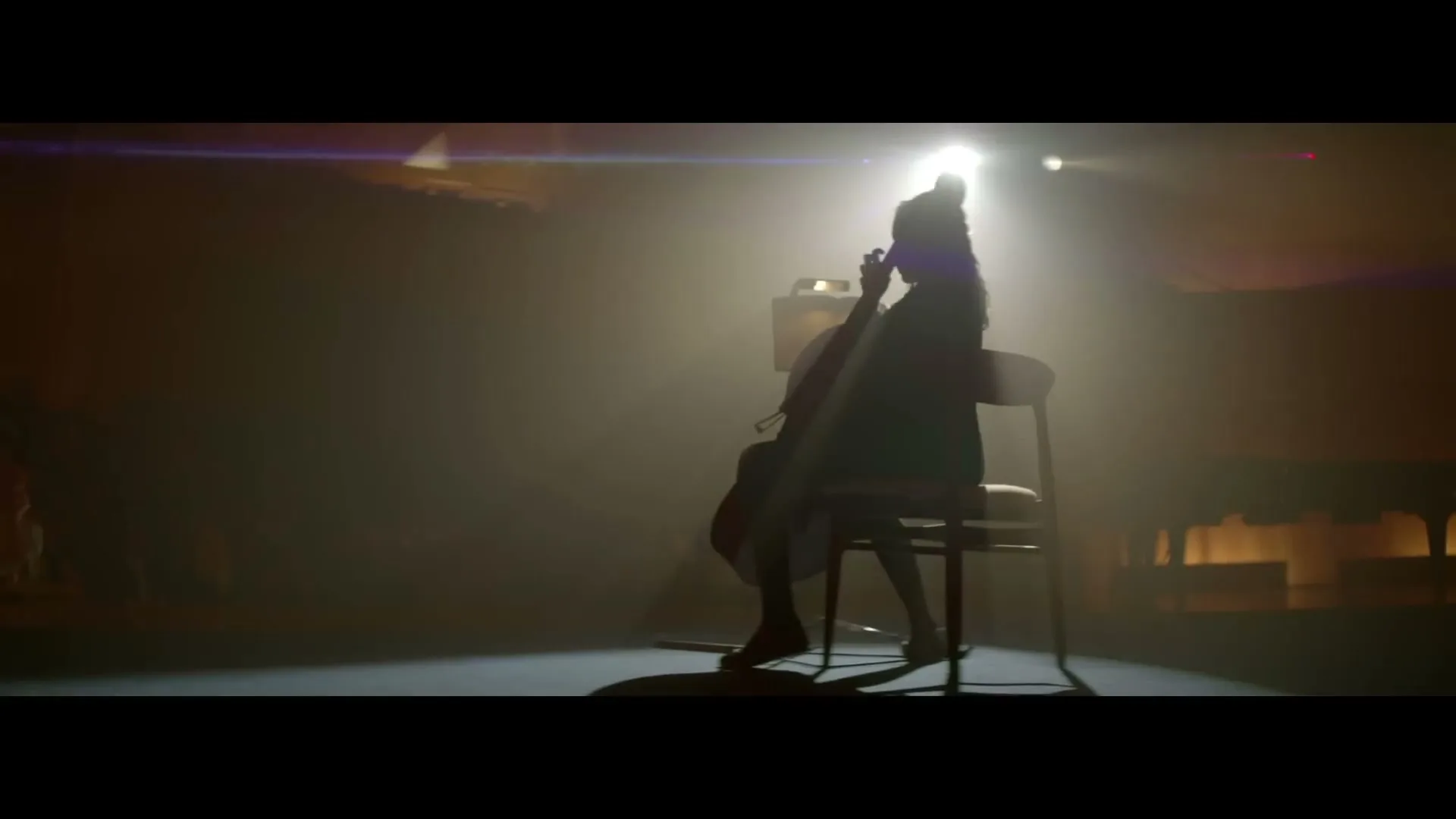 A young girl, silhouetted by a bright spotlight, sits on a chair playing a cello on a stage.