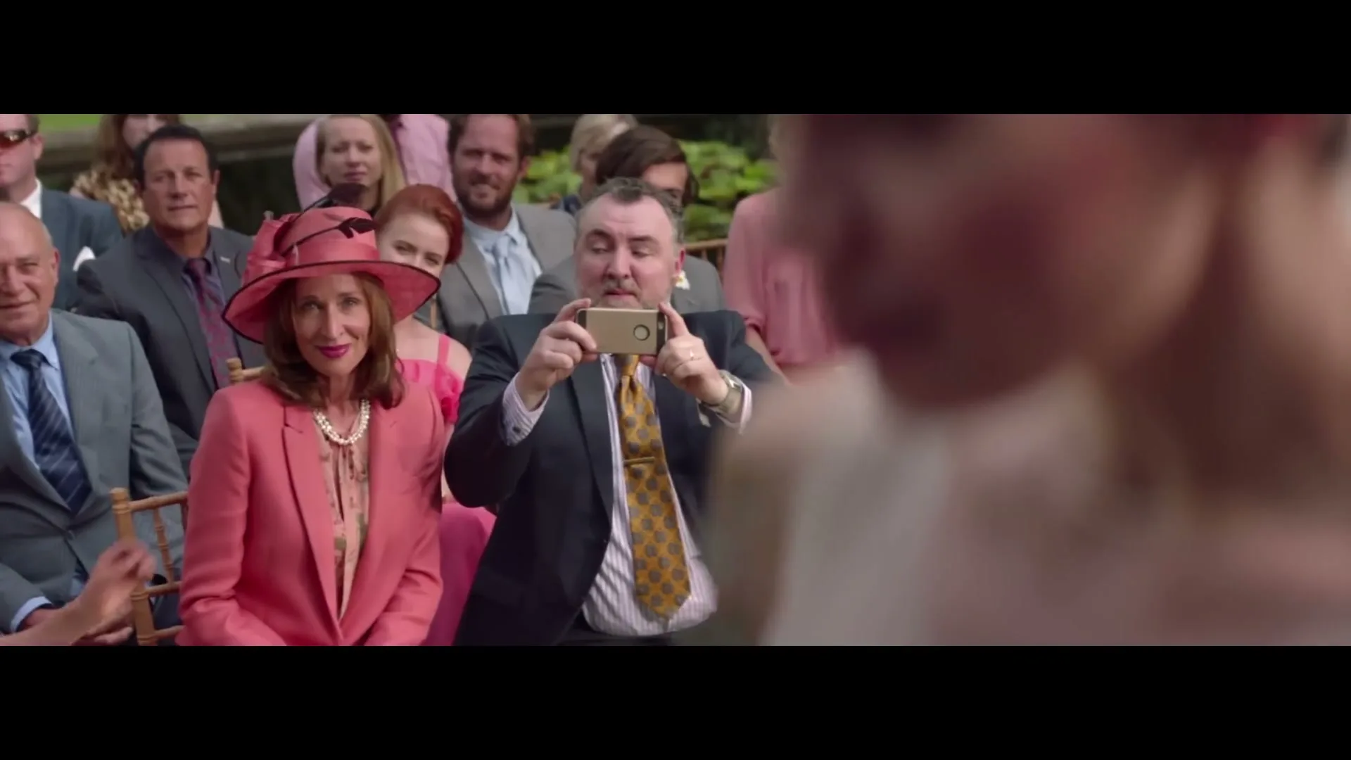 A man in a suit and tie holds up a smartphone to record a wedding, while a woman in a pink suit and hat sits beside him, smiling.