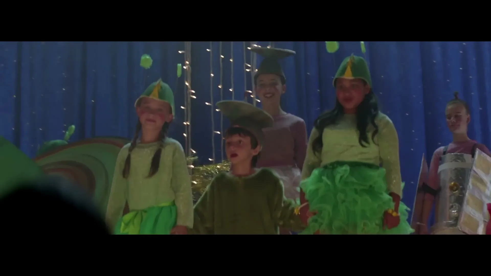 Four children in green alien costumes stand on a stage with blue curtains and string lights, looking up and smiling during a school play.