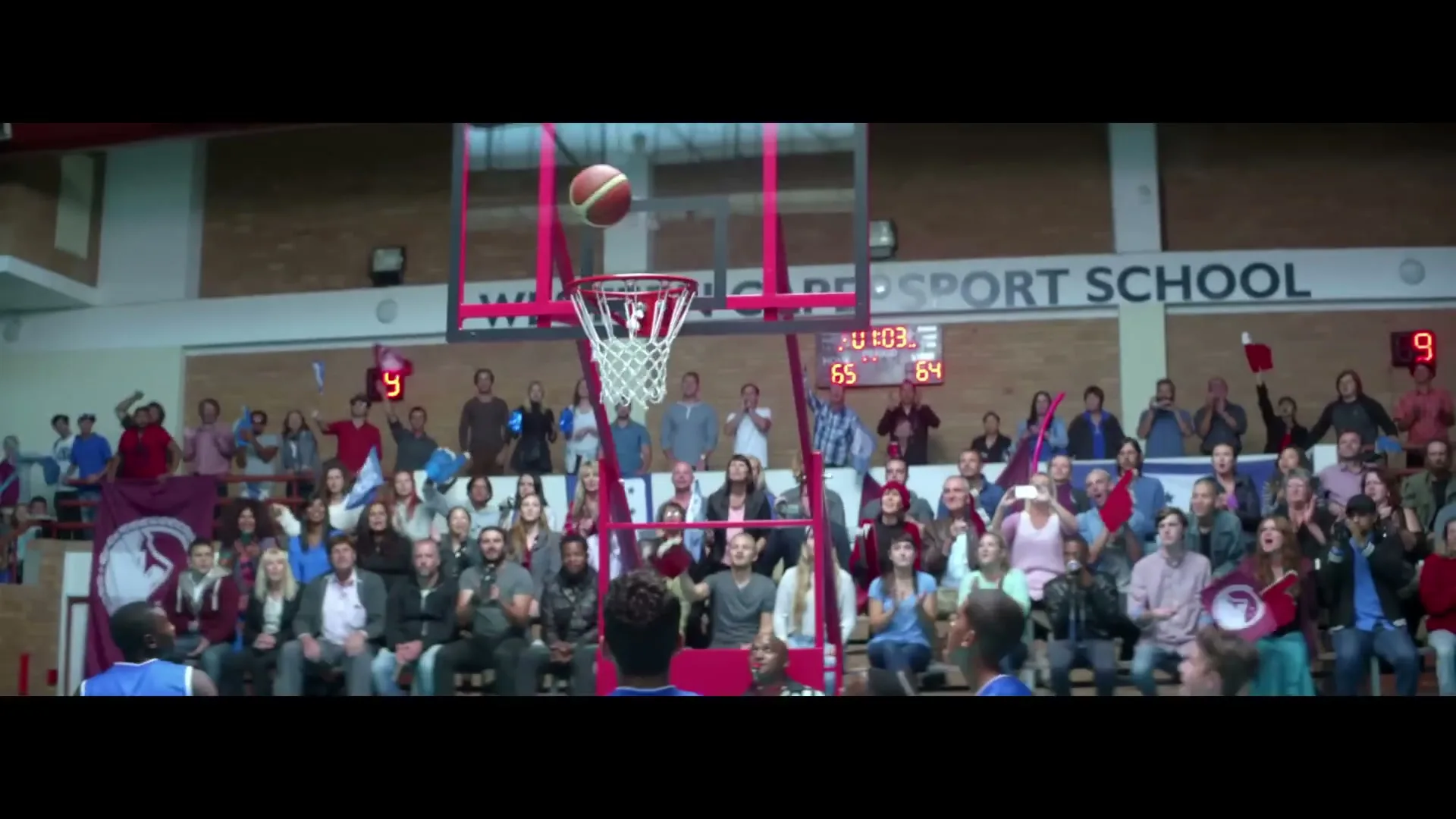 A basketball game is underway in a school gym, with a player shooting the ball towards the hoop as spectators watch from the stands.