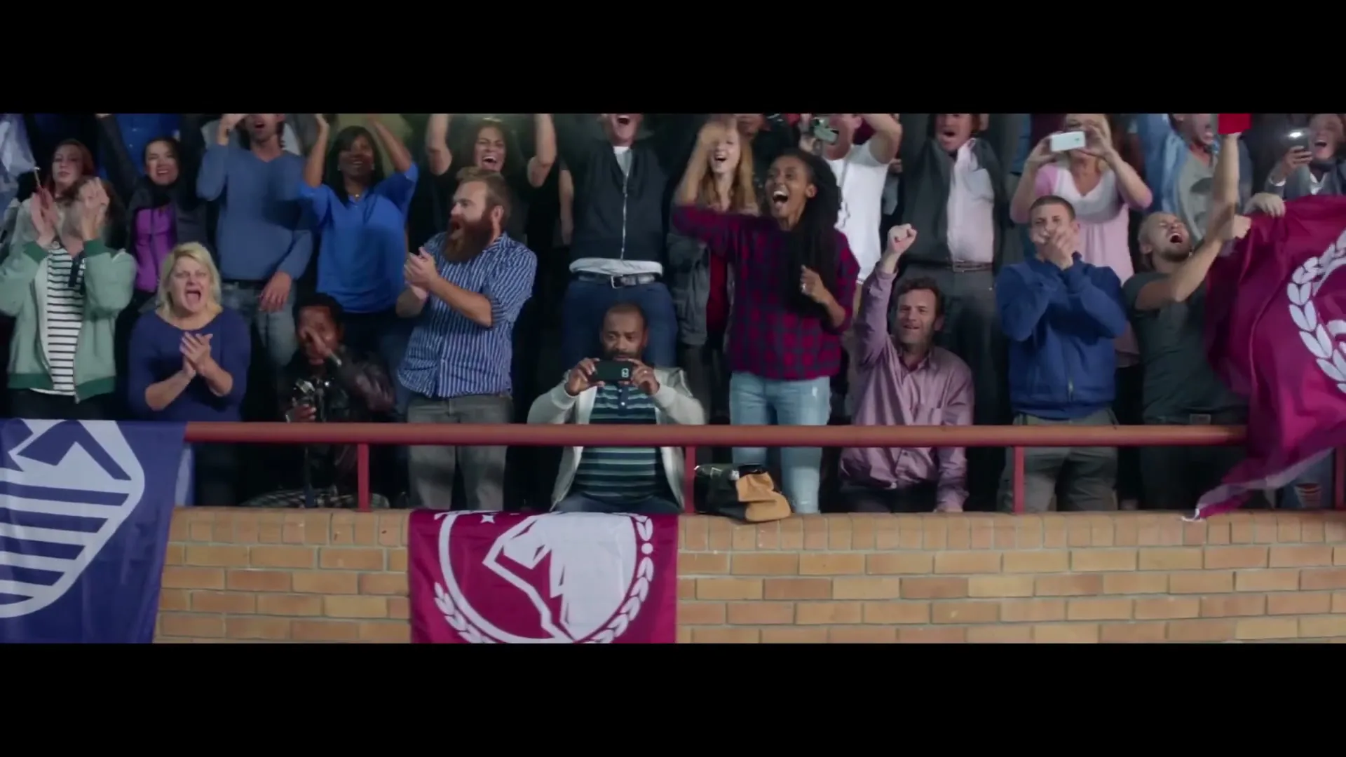 A diverse crowd of people in a stadium cheer and clap, with some holding up phones to record the event.