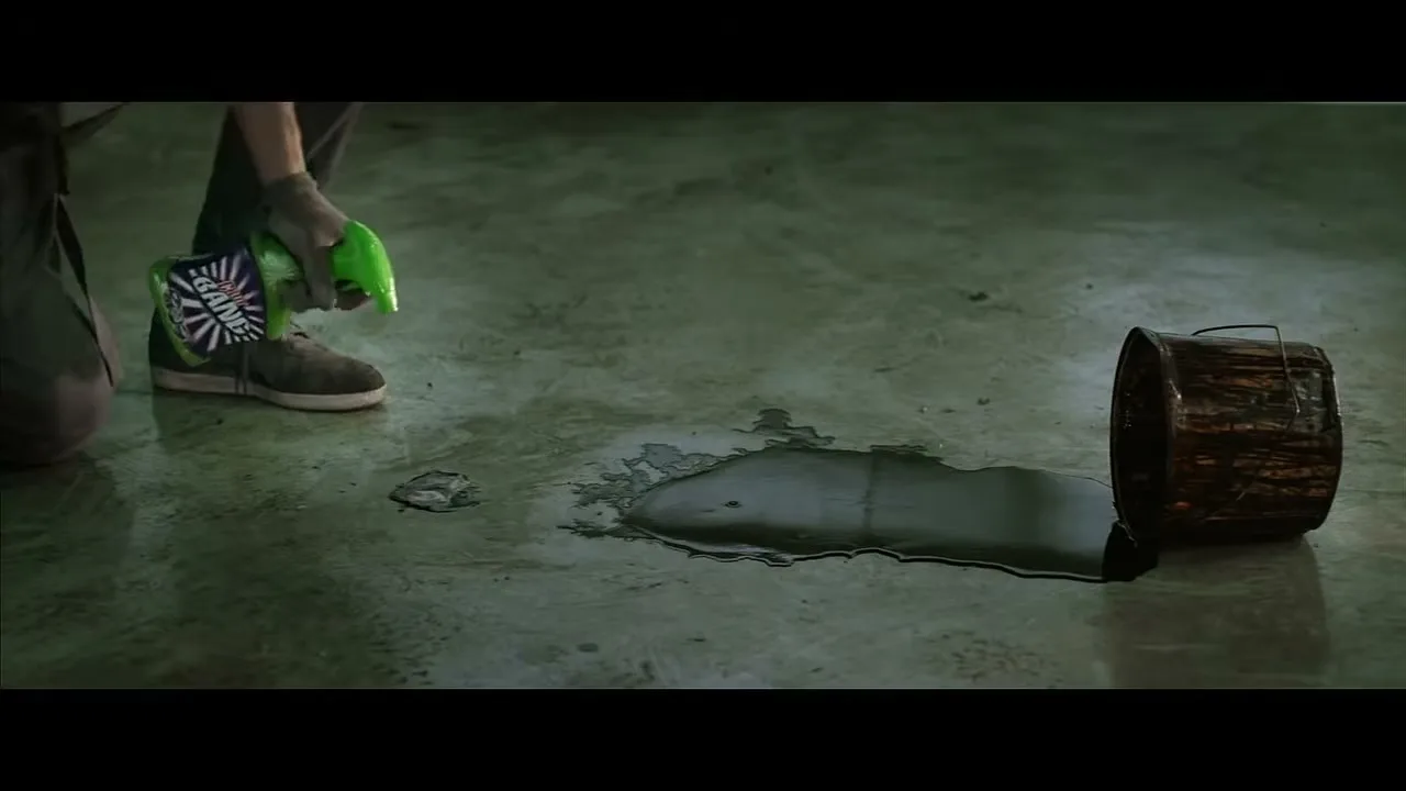 A person in work clothes and sneakers sprays a cleaning product on a dark liquid spill next to an overturned bucket on a concrete floor.