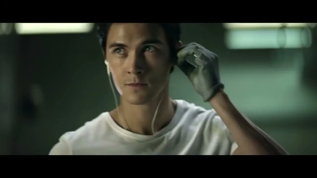 A young man in a white t-shirt and a grey glove listens to music through earbuds in a garage.