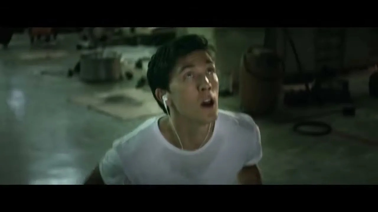 A young man in a white t-shirt and earbuds looks up with a surprised expression in a dimly lit industrial space.