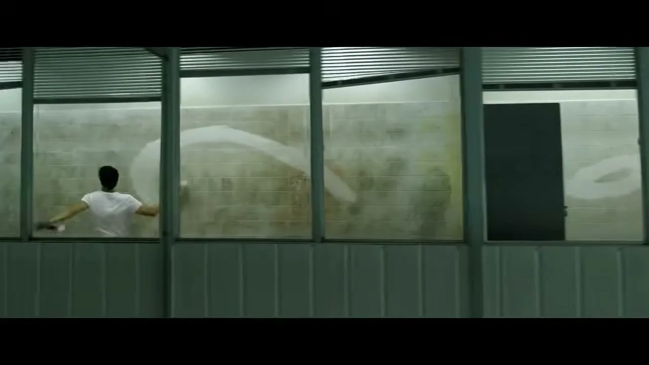 A man in a white t-shirt and dark pants cleans a large window with a squeegee, creating white streaks on the glass in an industrial setting.