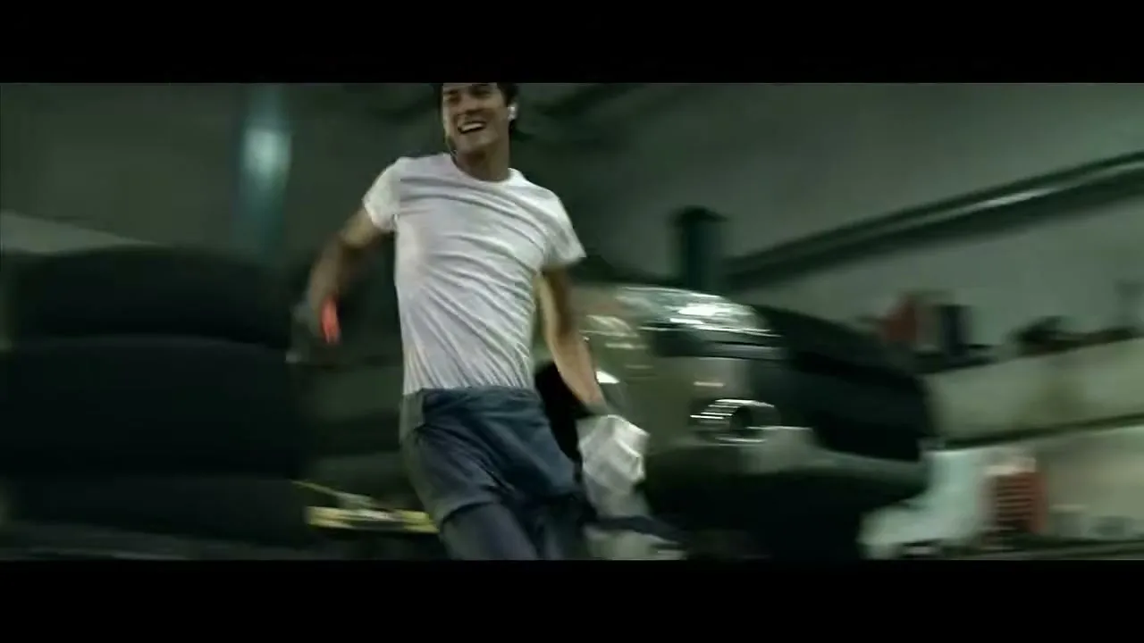 A young male mechanic in a white t-shirt and blue jeans dances energetically around a car on a lift in a messy garage, holding a cleaning product.