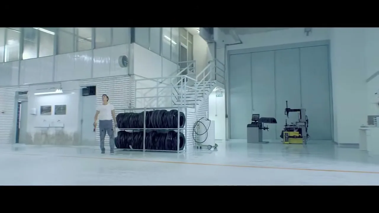 A male mechanic in a white t-shirt and grey pants walks across a clean, bright garage with stacks of tires and equipment.
