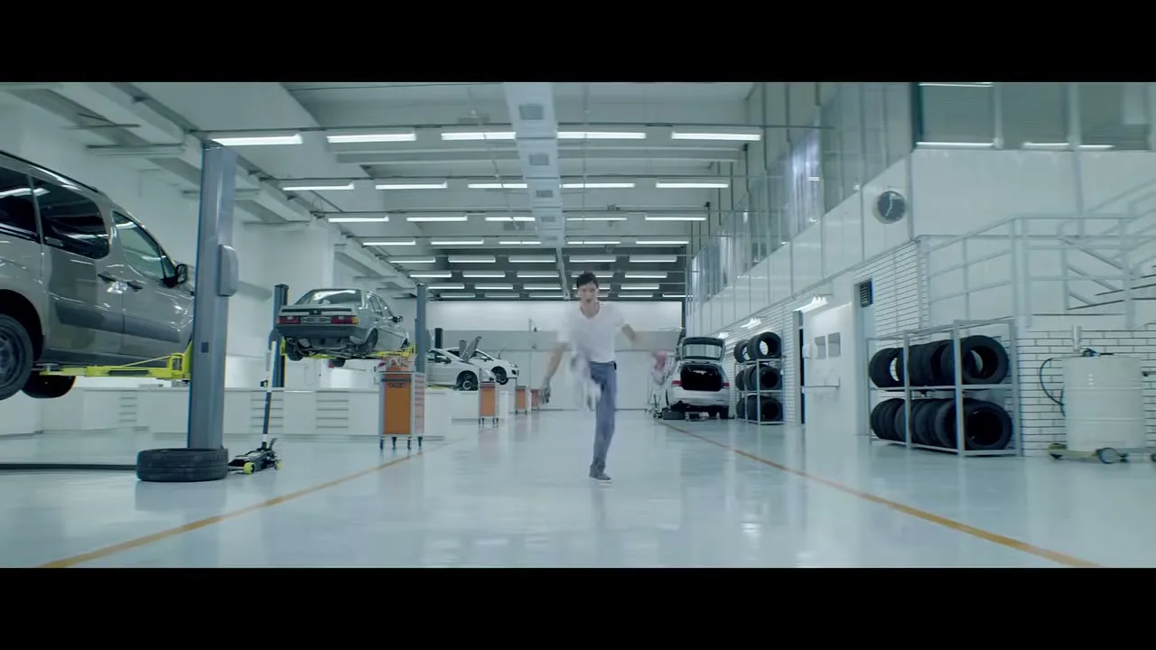 A young male mechanic in a white t-shirt and grey overalls dances energetically in a clean, brightly lit car repair shop.
