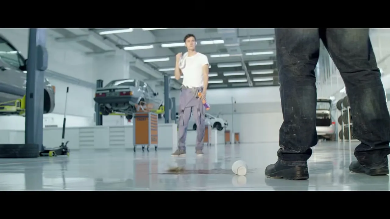 A mechanic in a white t-shirt and grey overalls stands in a clean garage, looking at a spilled cup of coffee on the floor.