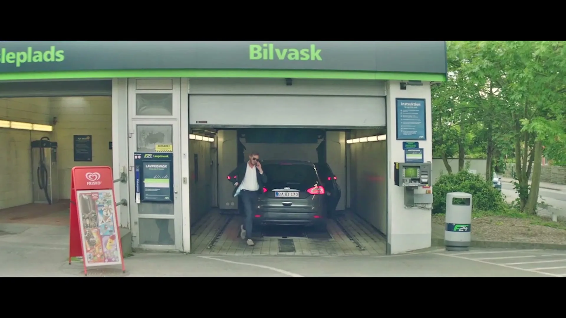 A man in a suit talks on the phone while walking out of a car wash, leaving his dark grey Ford car inside.