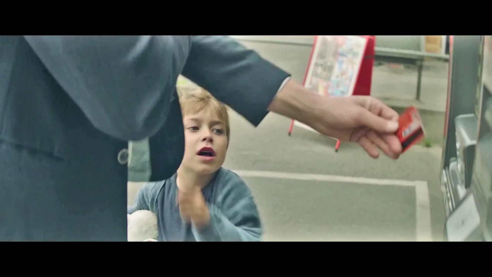 A young boy with blonde hair looks up at an adult's arm as they try to insert a card into a machine outdoors.