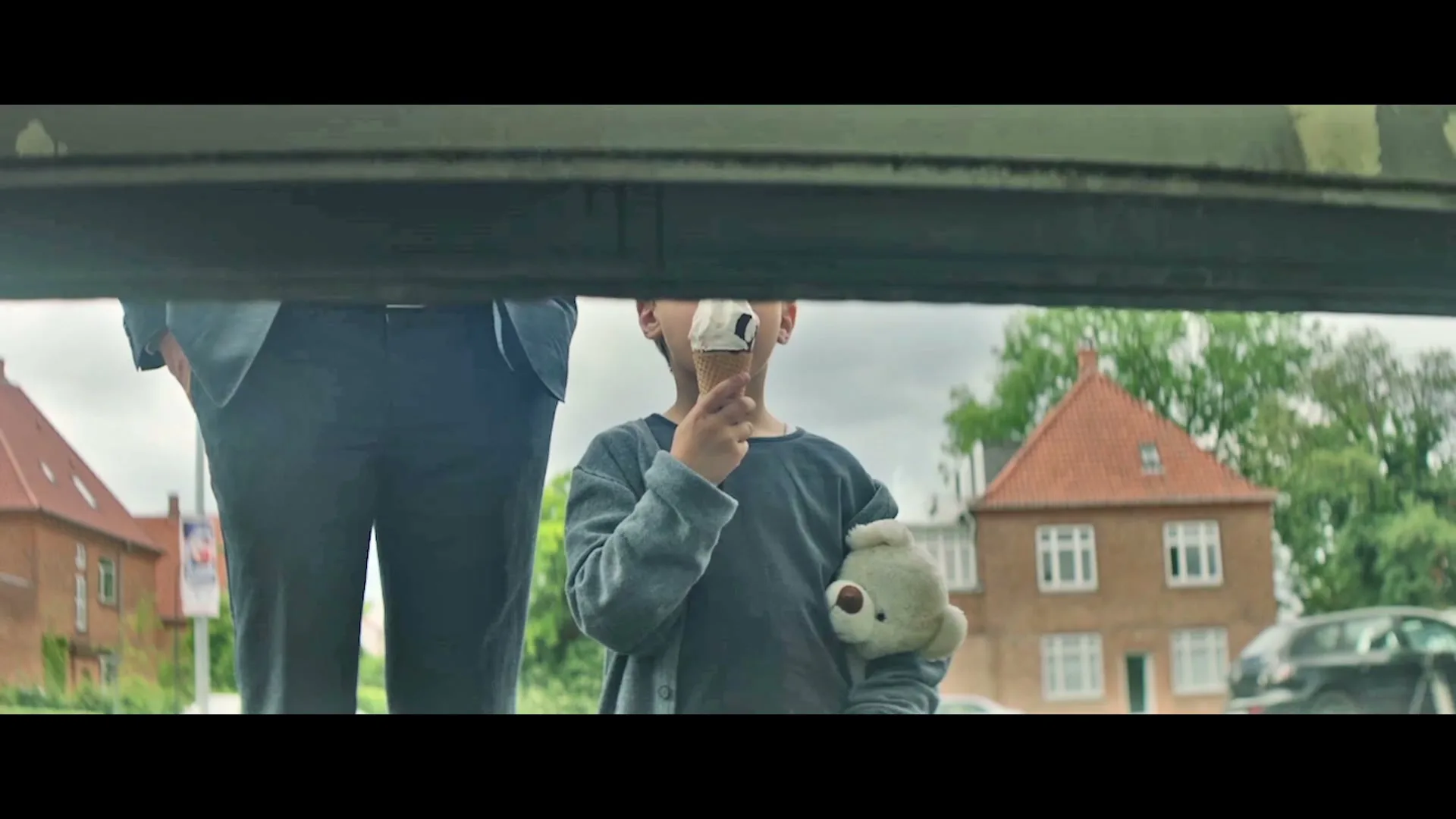 A young boy with messy hair, holding a teddy bear and an ice cream cone, stands next to an adult's legs as a garage door opens, revealing a suburban street.