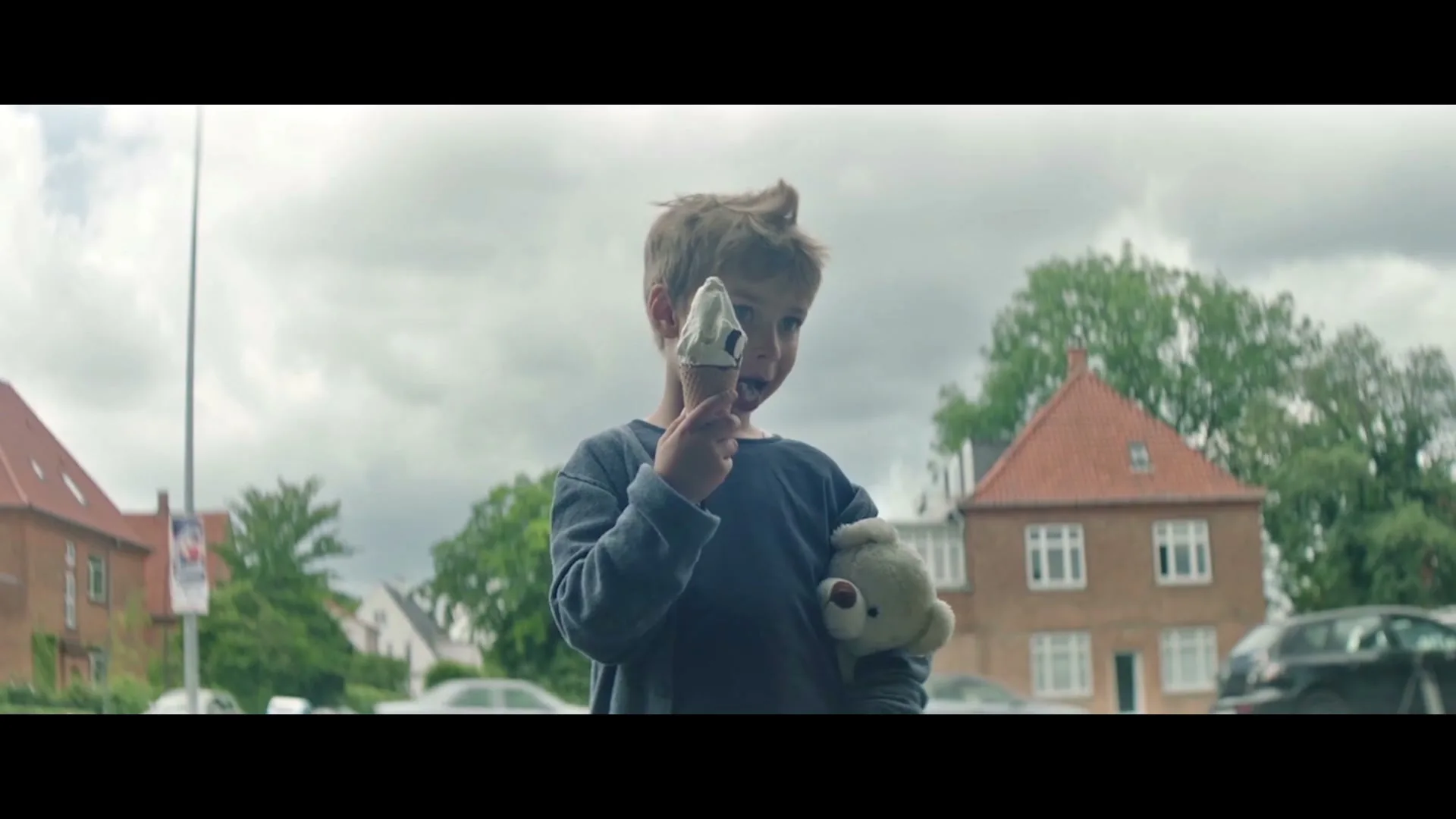 A young boy with spiky hair holds an ice cream cone to his face and a teddy bear in his arm, standing outdoors on a cloudy day.