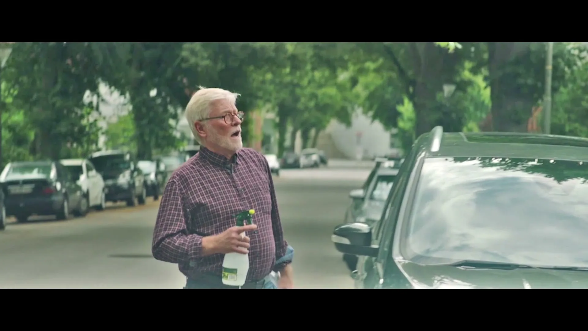 An older man with white hair and glasses, wearing a plaid shirt, holds a spray bottle and looks surprised next to a parked car on a tree-lined street.