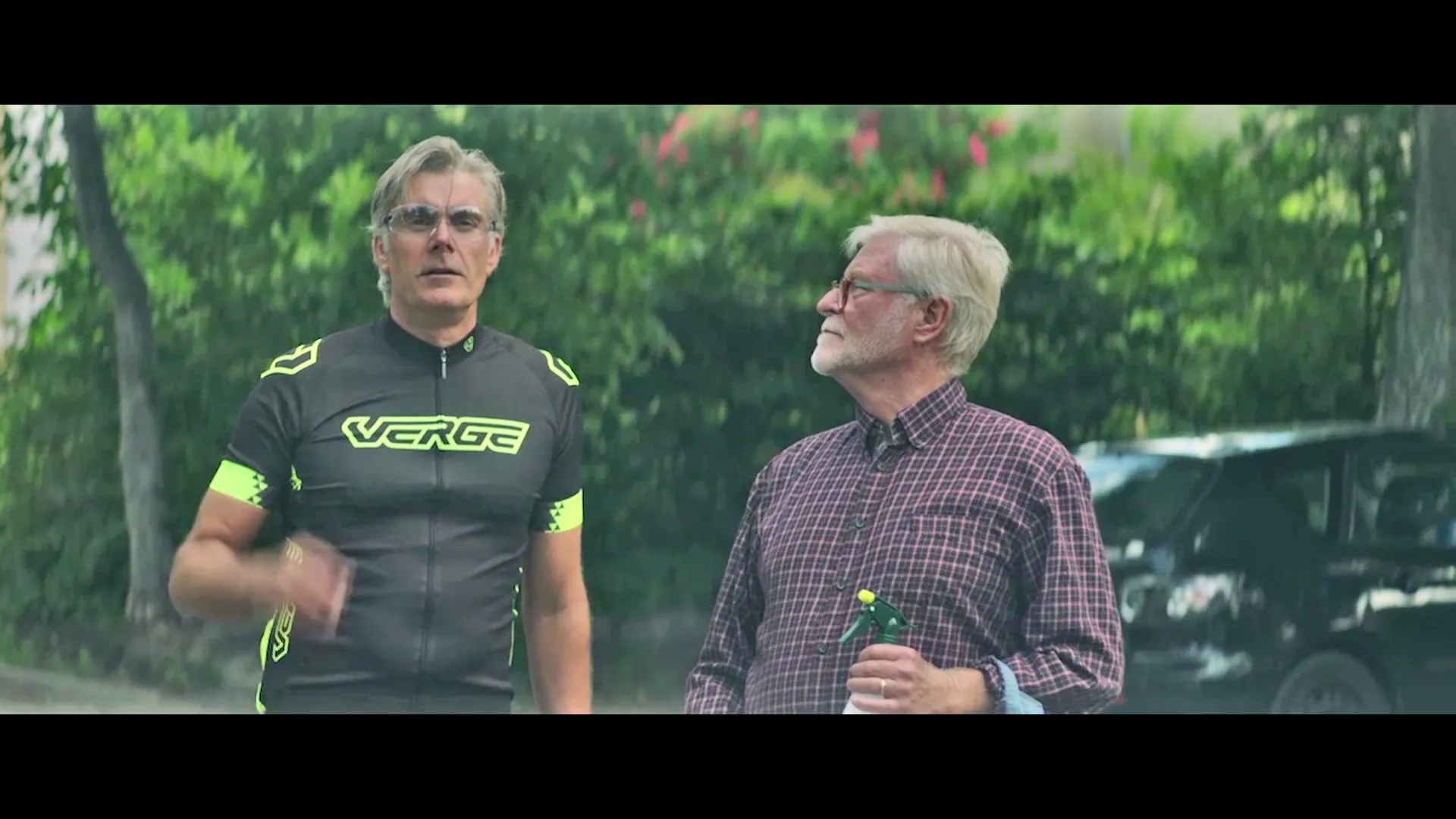 Two men, one in a cycling jersey and one in a plaid shirt holding a spray bottle, stand outside looking up at something off-screen.