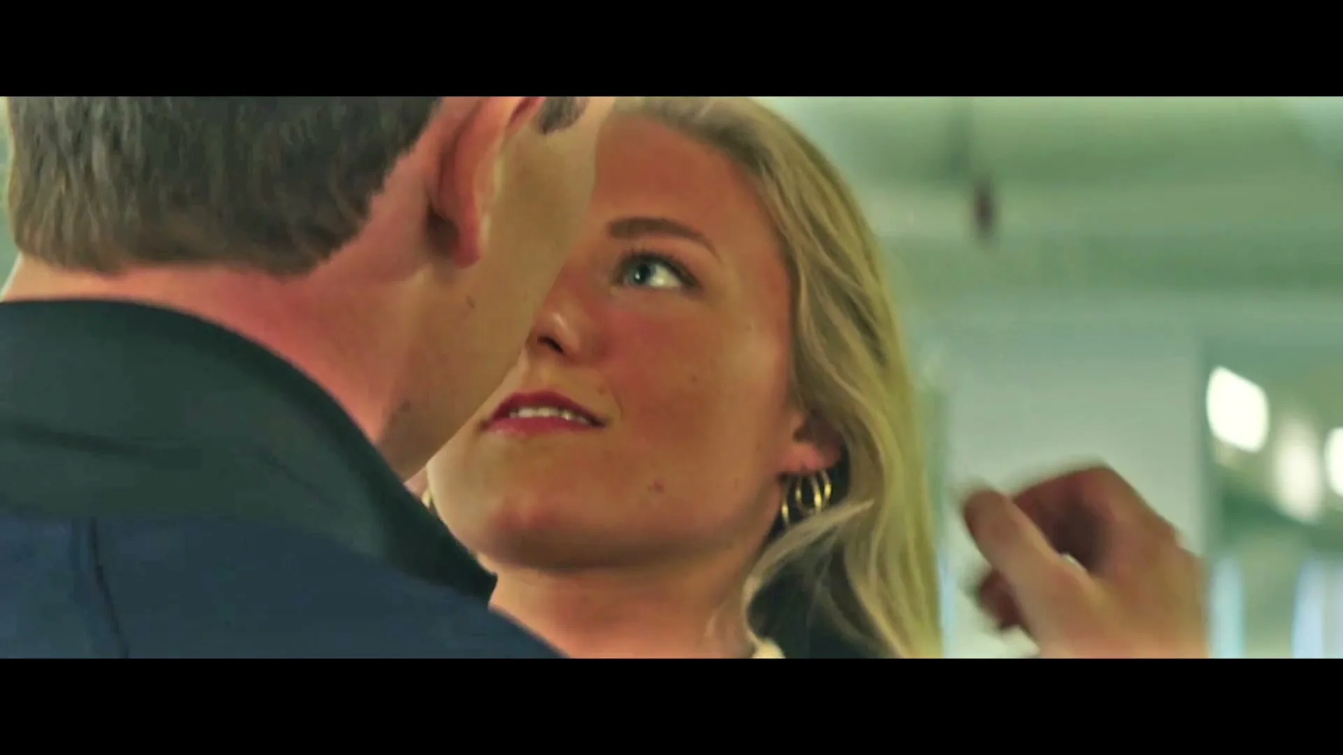 A young blonde woman with hoop earrings looks at a man, who then kisses her on the cheek in an interior setting.