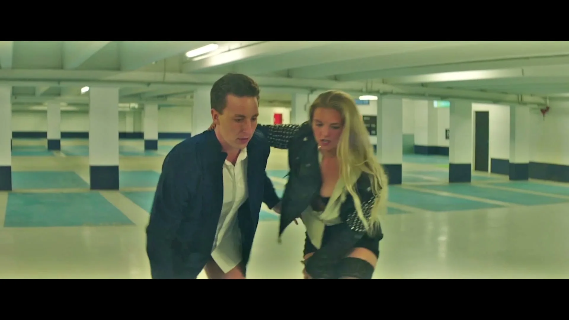 A young man and woman, dressed in casual attire, are seen stumbling and falling in an empty, brightly lit parking garage.