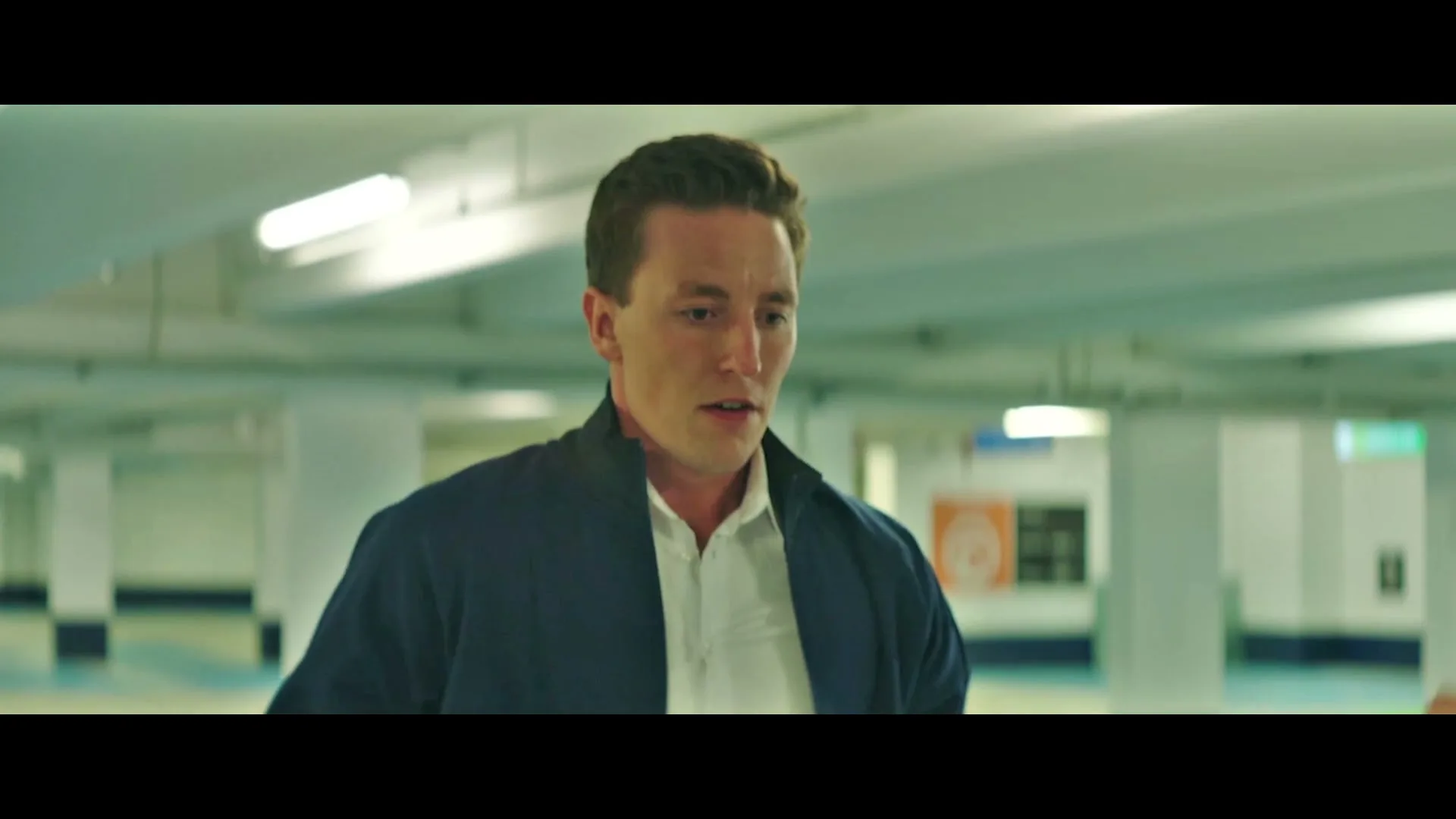 A young man in a blue jacket and white shirt looks down and then up, talking in an underground parking garage.