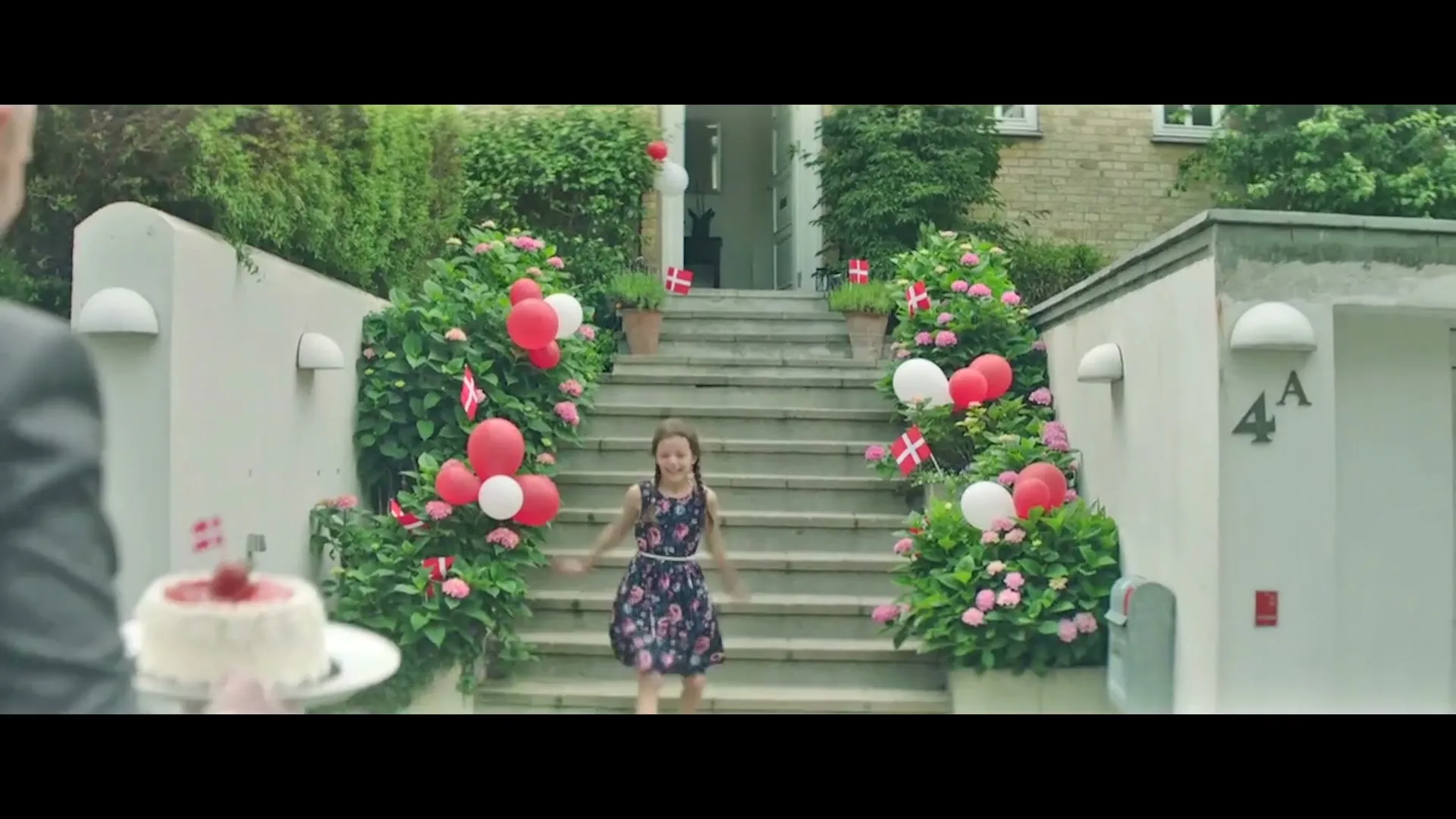 A young girl in a floral dress runs down outdoor stairs decorated with red and white balloons and Danish flags, as a man approaches with a cake.