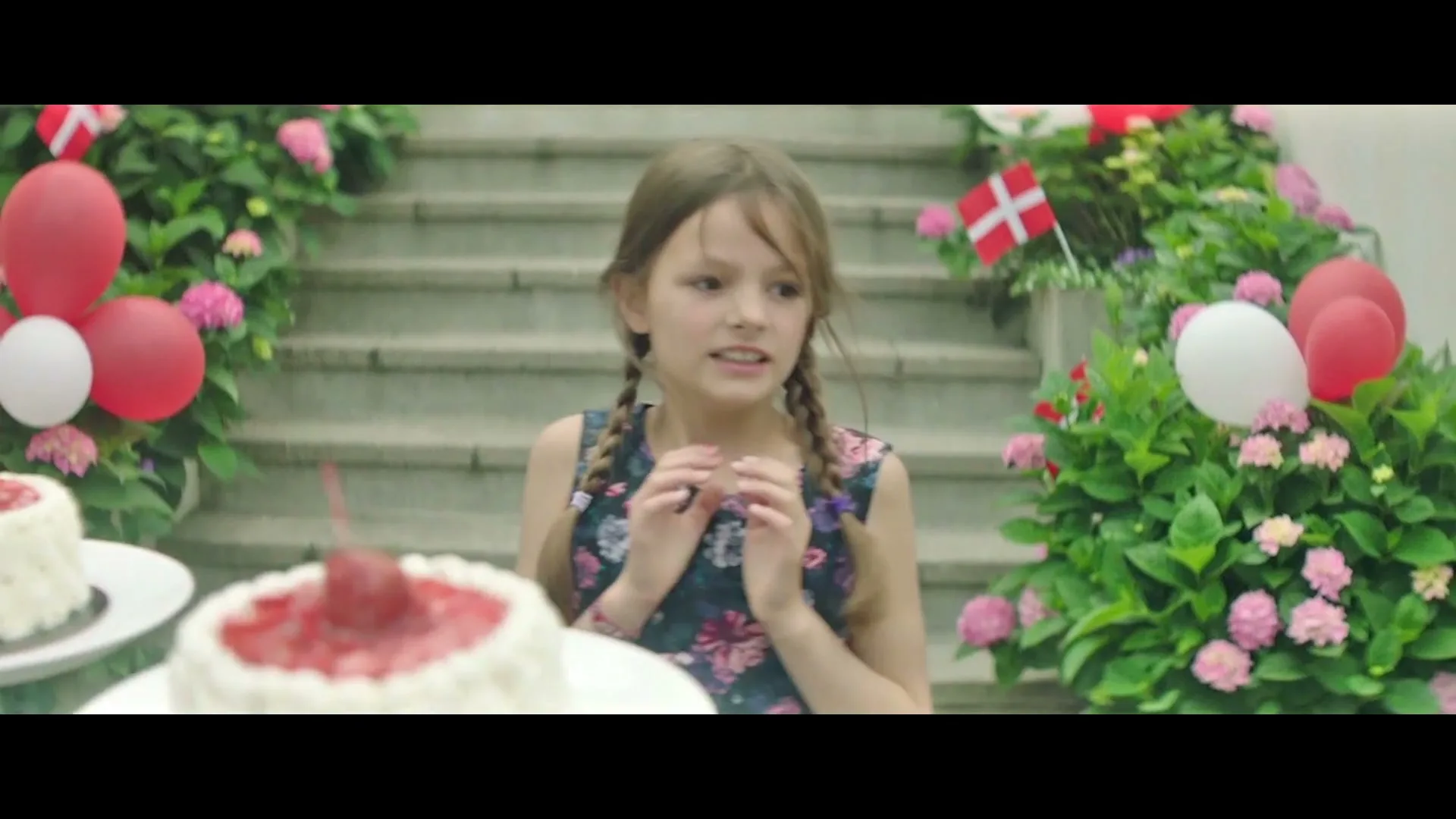 A young girl with pigtails in a floral dress claps her hands, then looks down at two strawberry cakes on a table in a garden.