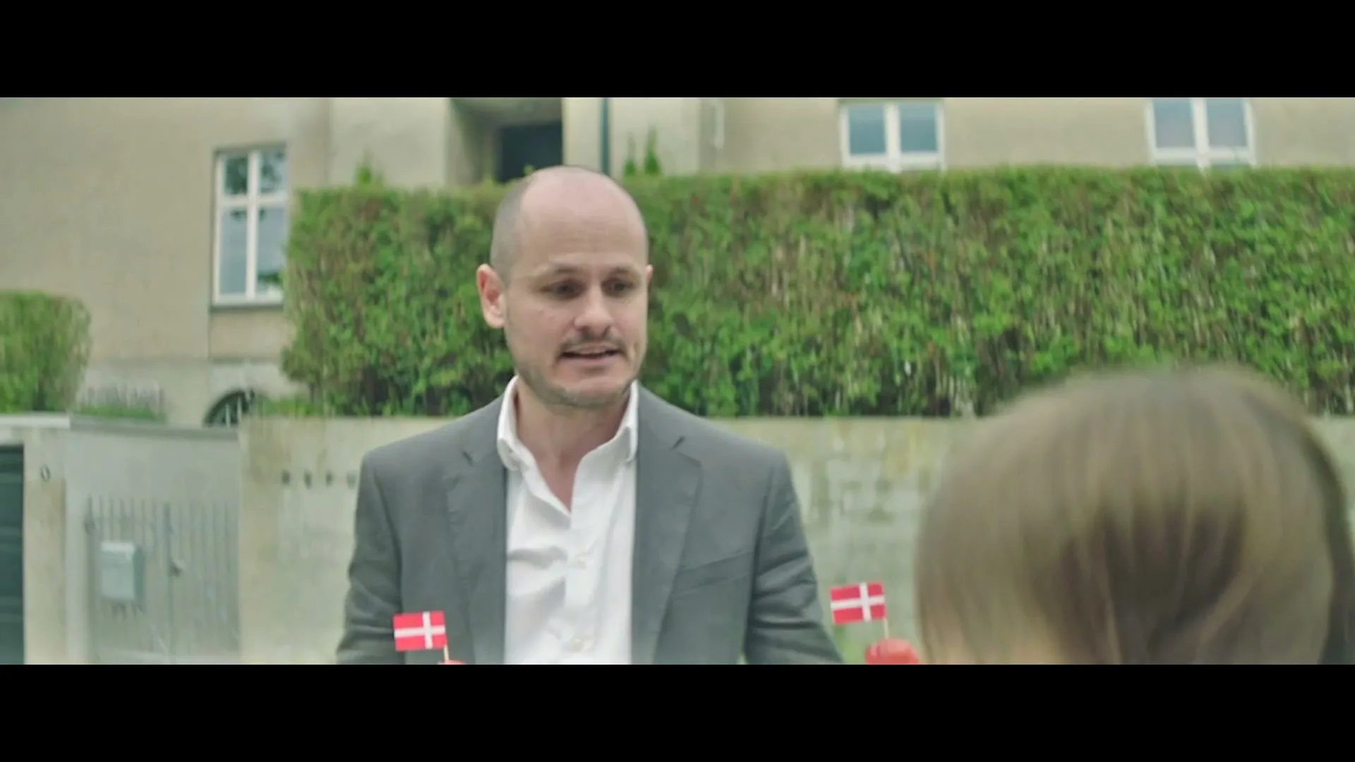 A bald man in a suit with Danish flags pinned to his lapels looks at a child from the perspective of a car, on a suburban street.
