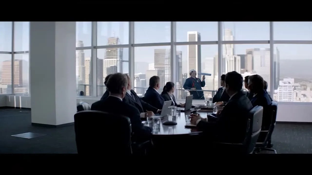 A group of business people in suits sit around a conference table in a high-rise office, looking at a window washer outside.