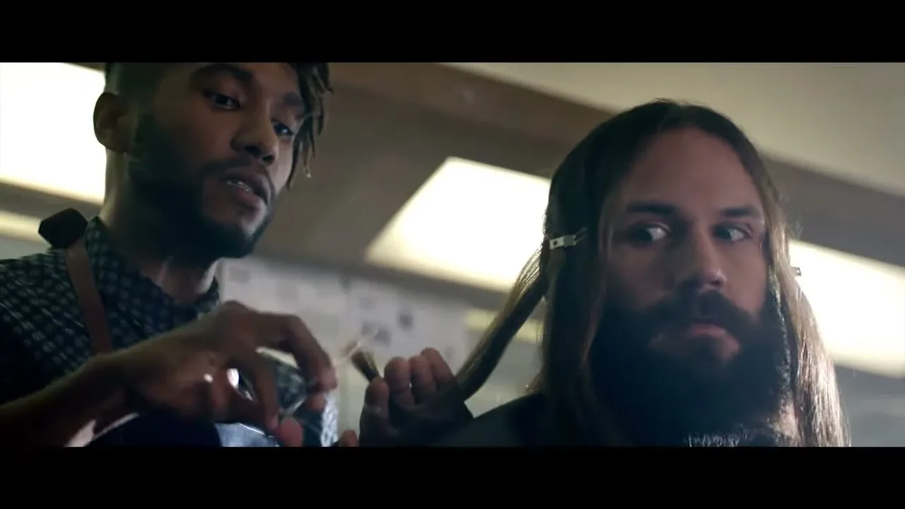 A male barber with dreadlocks cuts the long hair of a male client with a beard, who looks away nervously in a barbershop.