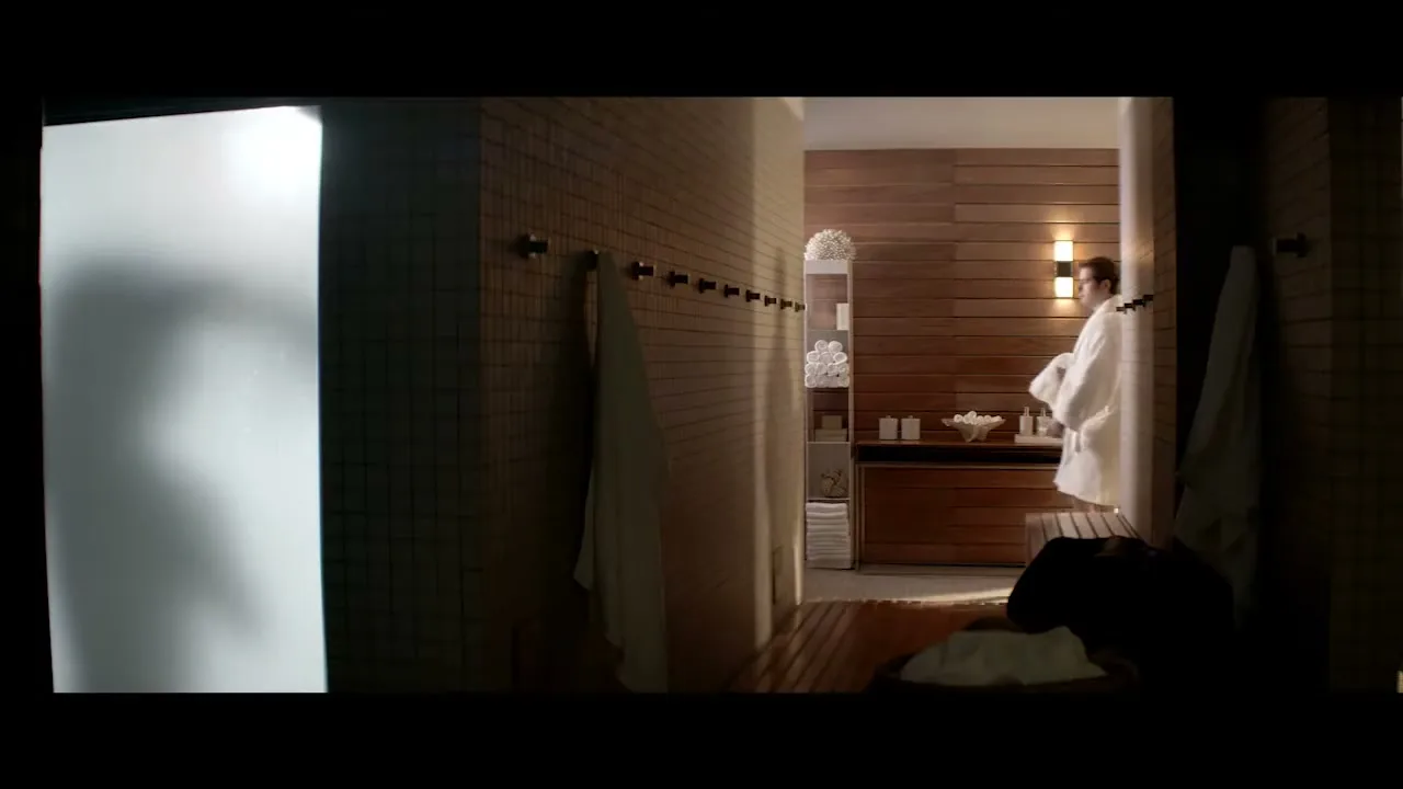 A man in a white robe walks out of a shower and into a luxurious spa changing room, with a frosted glass door on the left.