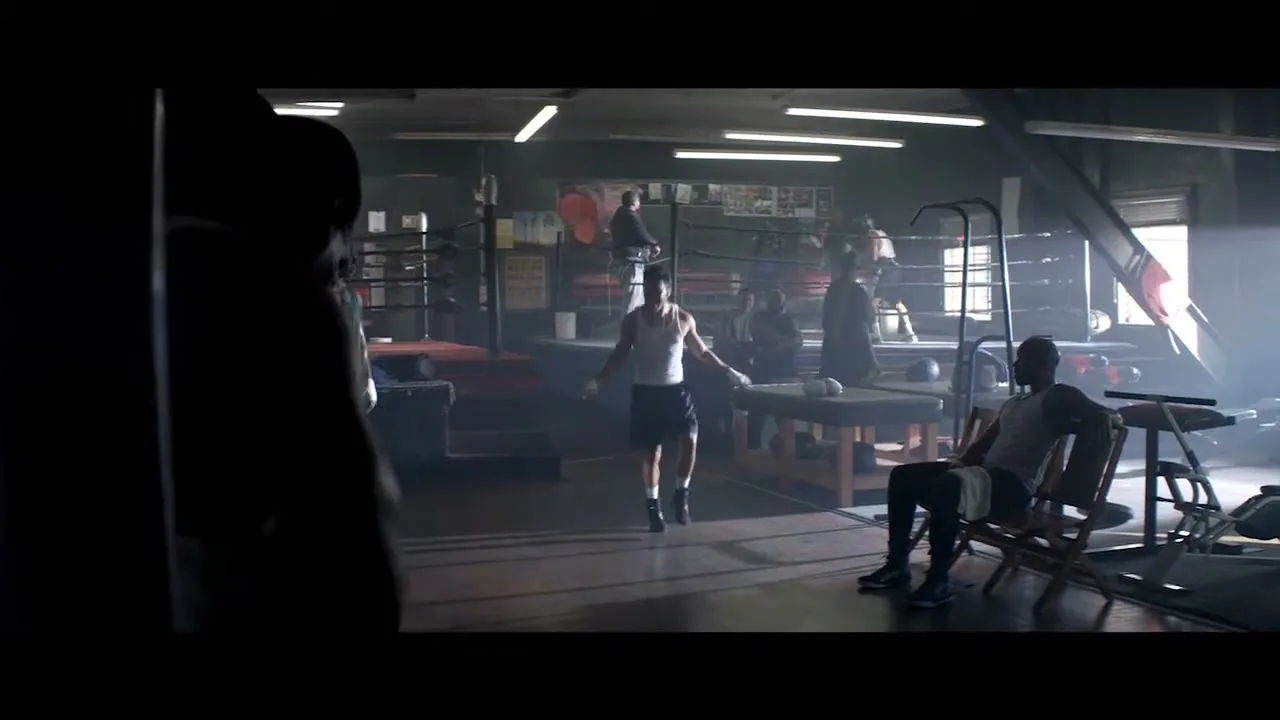 A man in a white tank top and black shorts jumps rope in the middle of a dimly lit boxing gym, while other men train around him.