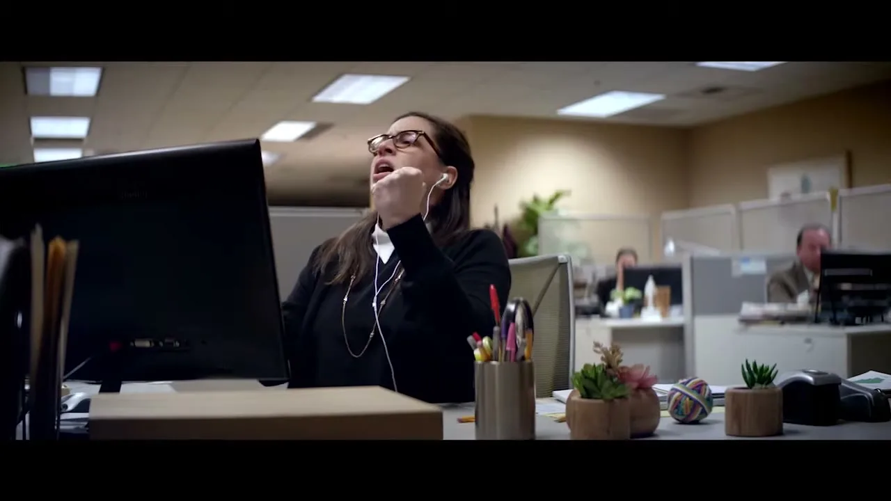 A woman in glasses and a black cardigan sings passionately into her computer screen while wearing earbuds in a cubicle office.
