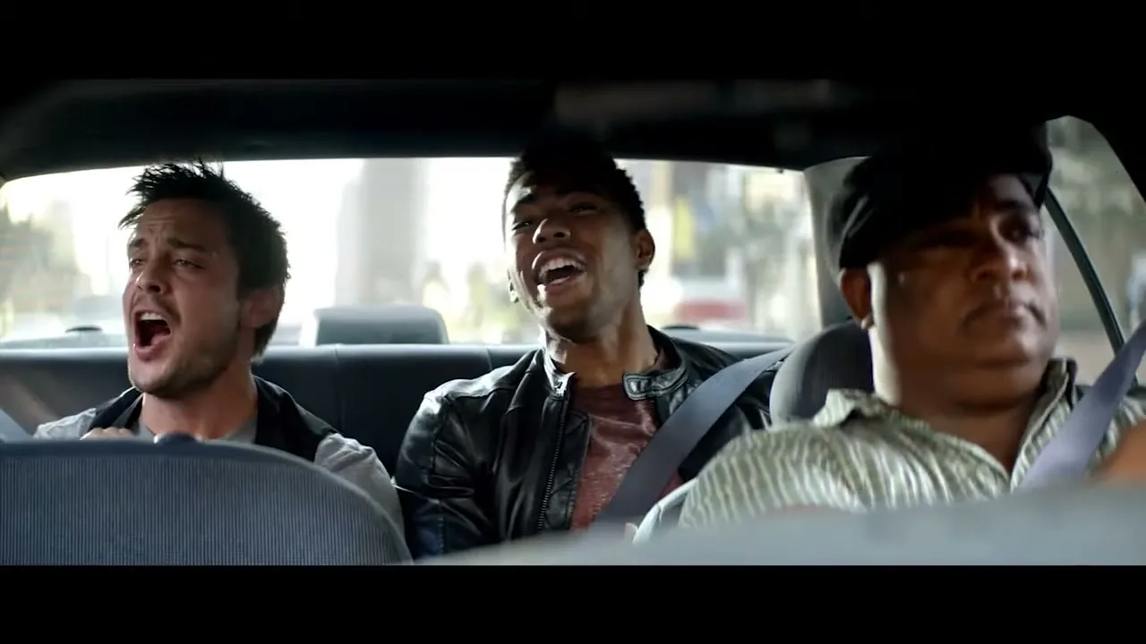 Three men in a taxi sing along to a song, with the man in the middle gesturing with his hand.