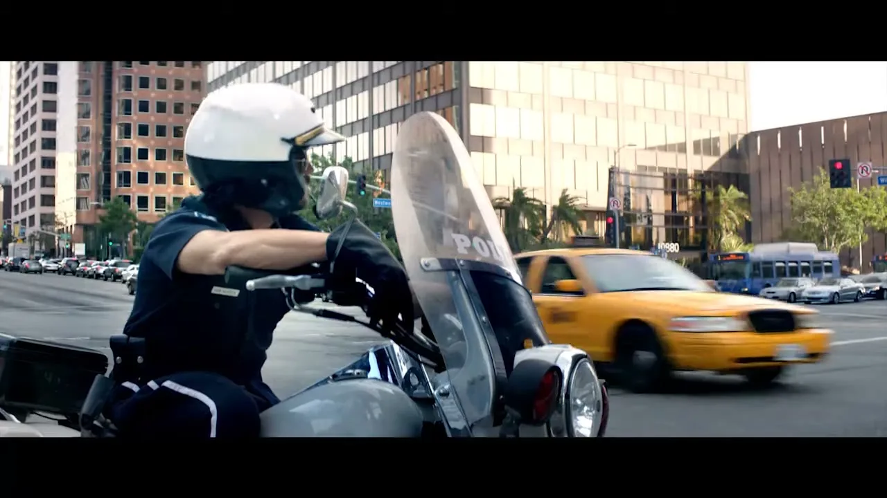 A male police officer in a white helmet and sunglasses sits on a motorcycle at an intersection in a city during the day.