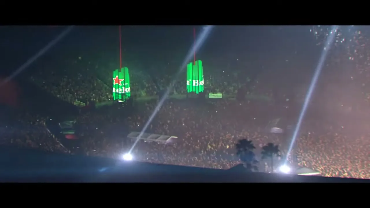 An extreme wide shot shows a large crowd at a night concert, with two large Heineken signs hanging above the stage, illuminated by spotlights.