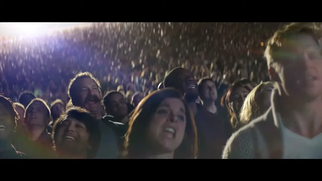 A diverse crowd of people, including a woman with dark hair and a man with a mustache, sing along at a concert in a large stadium.