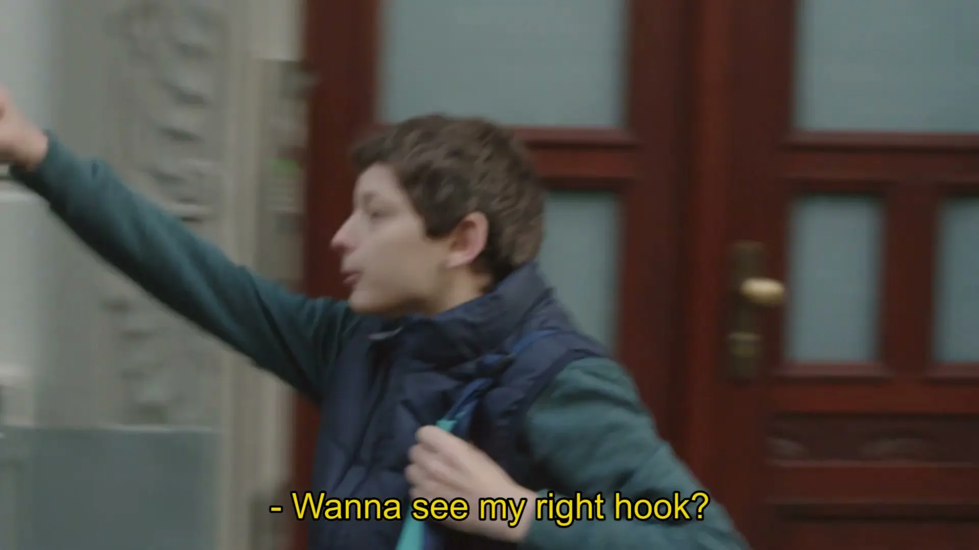 A man with a beard and a boy with curly hair are walking and talking on a city street, with the man playfully interacting with the boy.