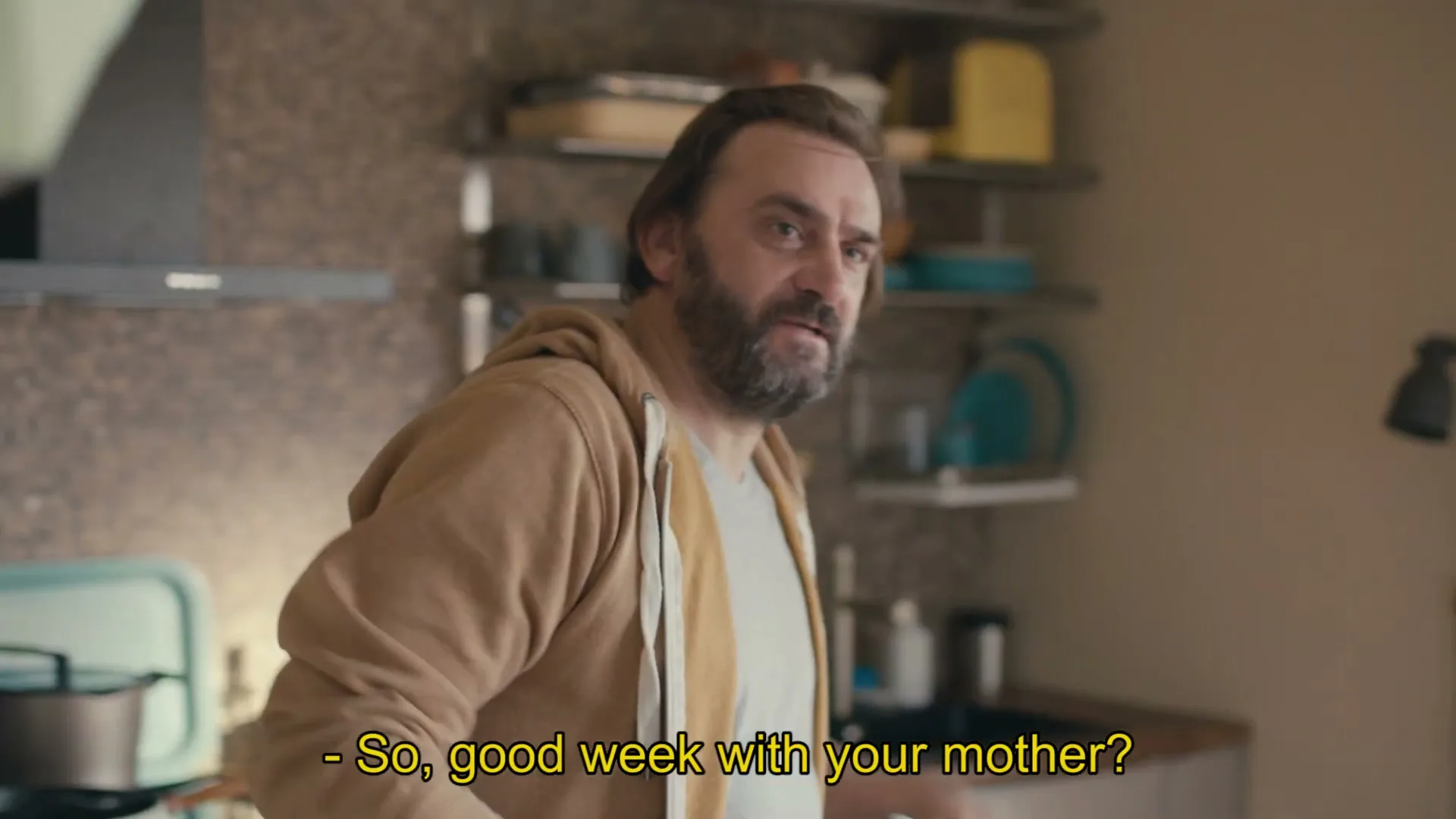 A middle-aged man with a beard, wearing a brown hoodie, looks over his shoulder and talks in a kitchen.