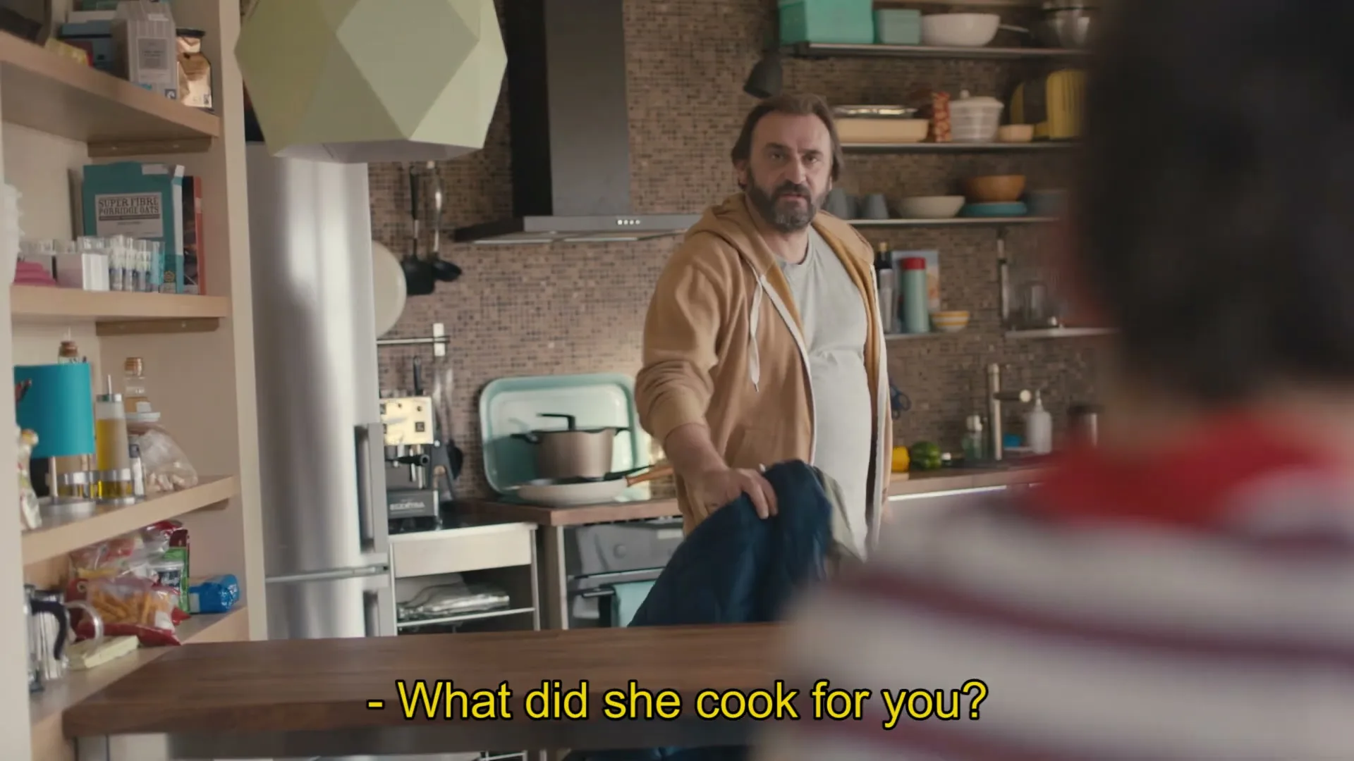 A man in a brown hoodie and grey t-shirt stands in a modern kitchen, looking at a child whose back is to the camera.