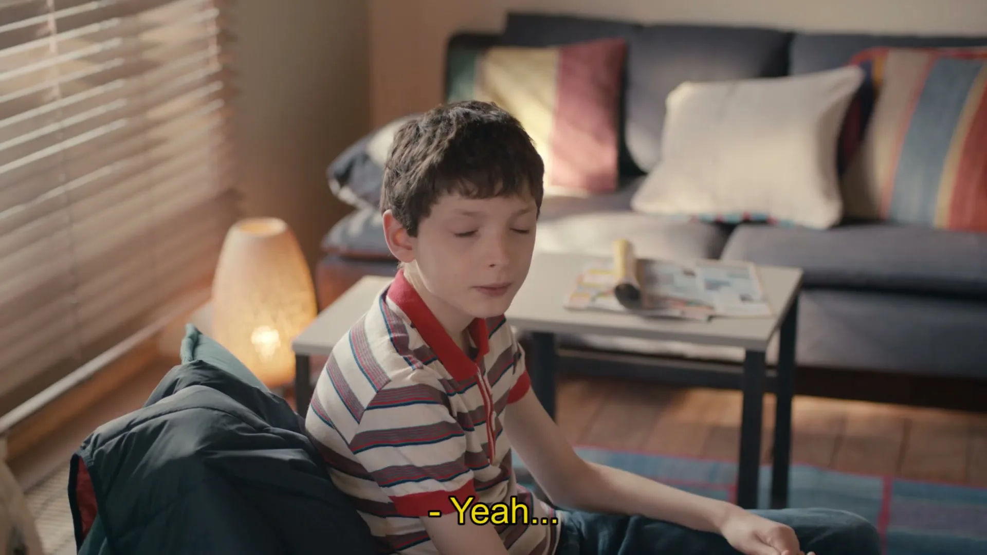 A young boy with dark hair and a striped shirt sits on a couch, looking off-camera with a thoughtful expression in a cozy living room.