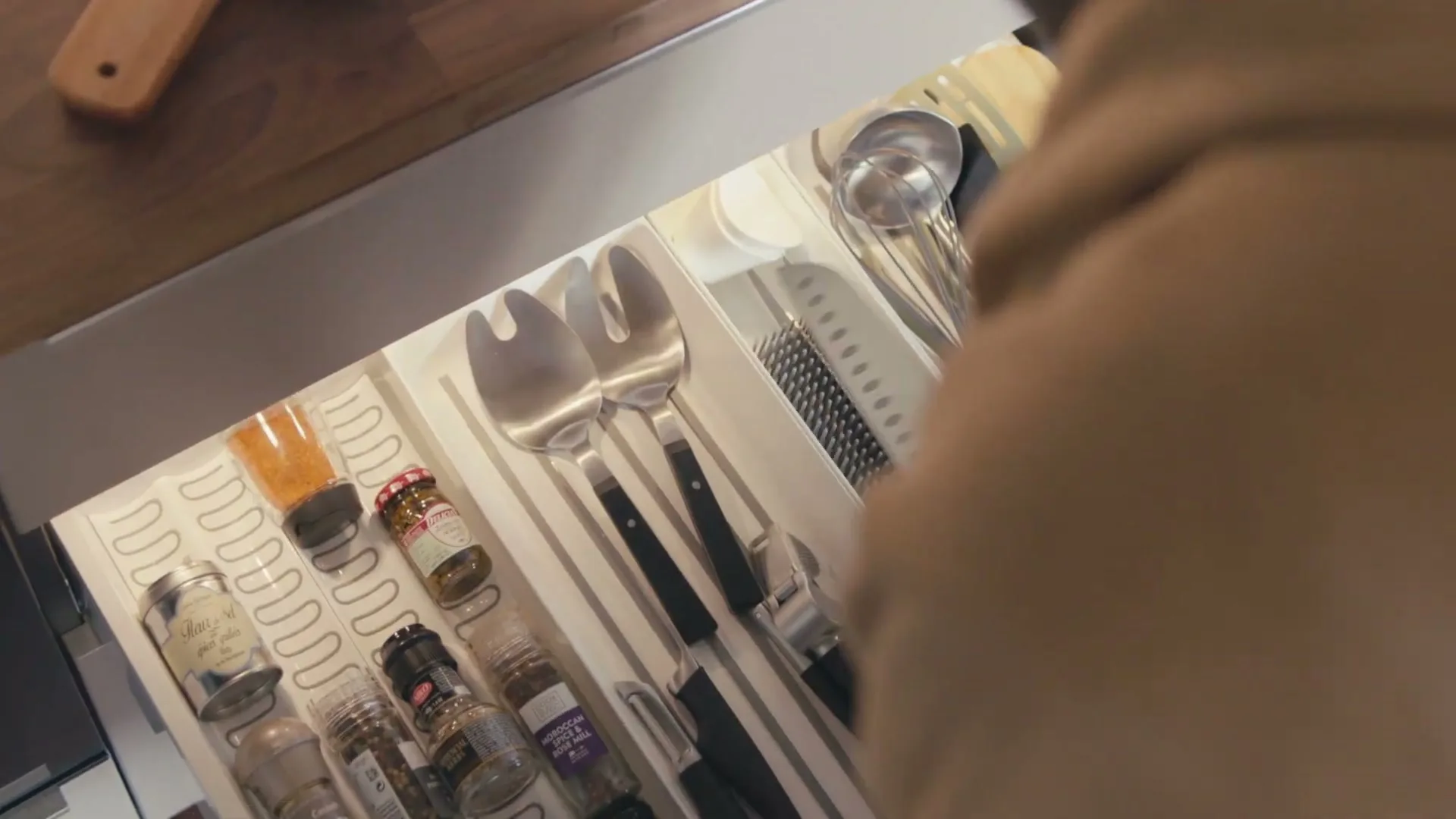 A person's hands open a kitchen drawer filled with utensils and spices, then close it, in a bright kitchen.