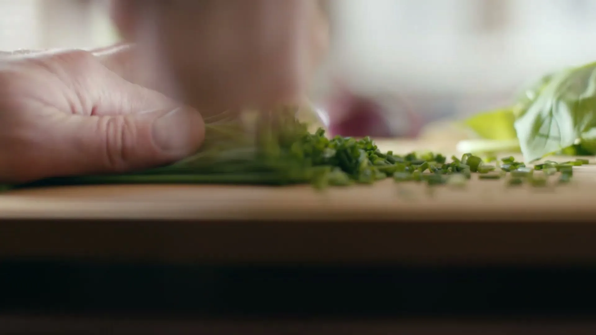 A person's hands are seen chopping chives on a wooden cutting board in a kitchen.