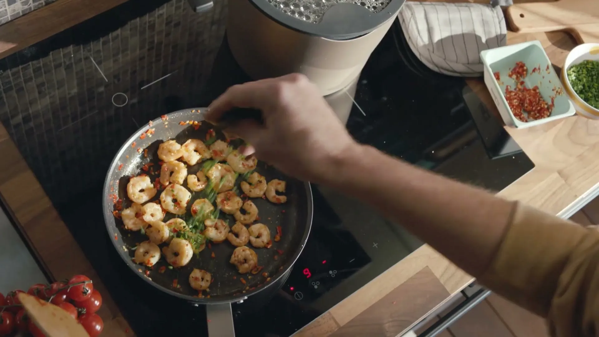 A person's arm sprinkles green herbs into a frying pan full of cooked shrimp on a black induction stovetop in a kitchen.