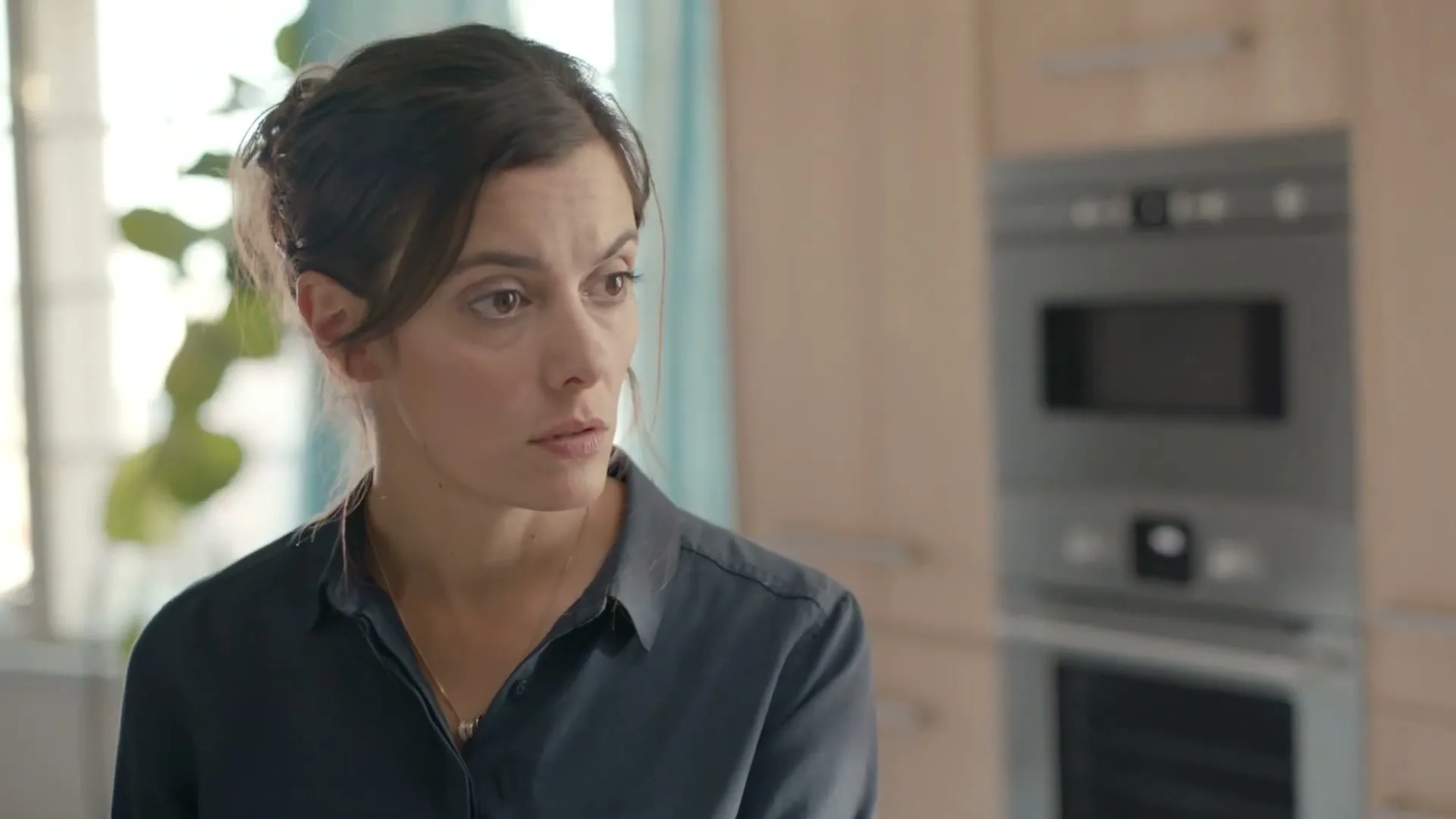 A woman with dark hair in a dark blue shirt looks to the right with a concerned expression in a modern kitchen.