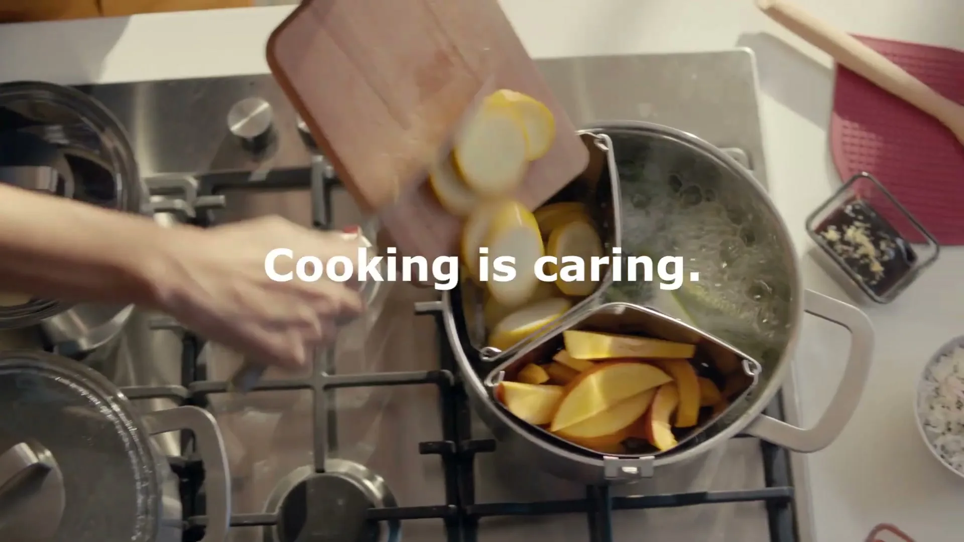 A person's hand slides sliced yellow squash from a wooden cutting board into a segmented pot on a stovetop, with the text "Cooking is caring." overlaid.