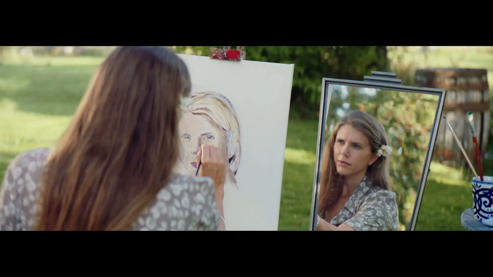 A young woman with long brown hair, wearing a floral dress, paints a self-portrait on an easel outdoors in a sunny garden.