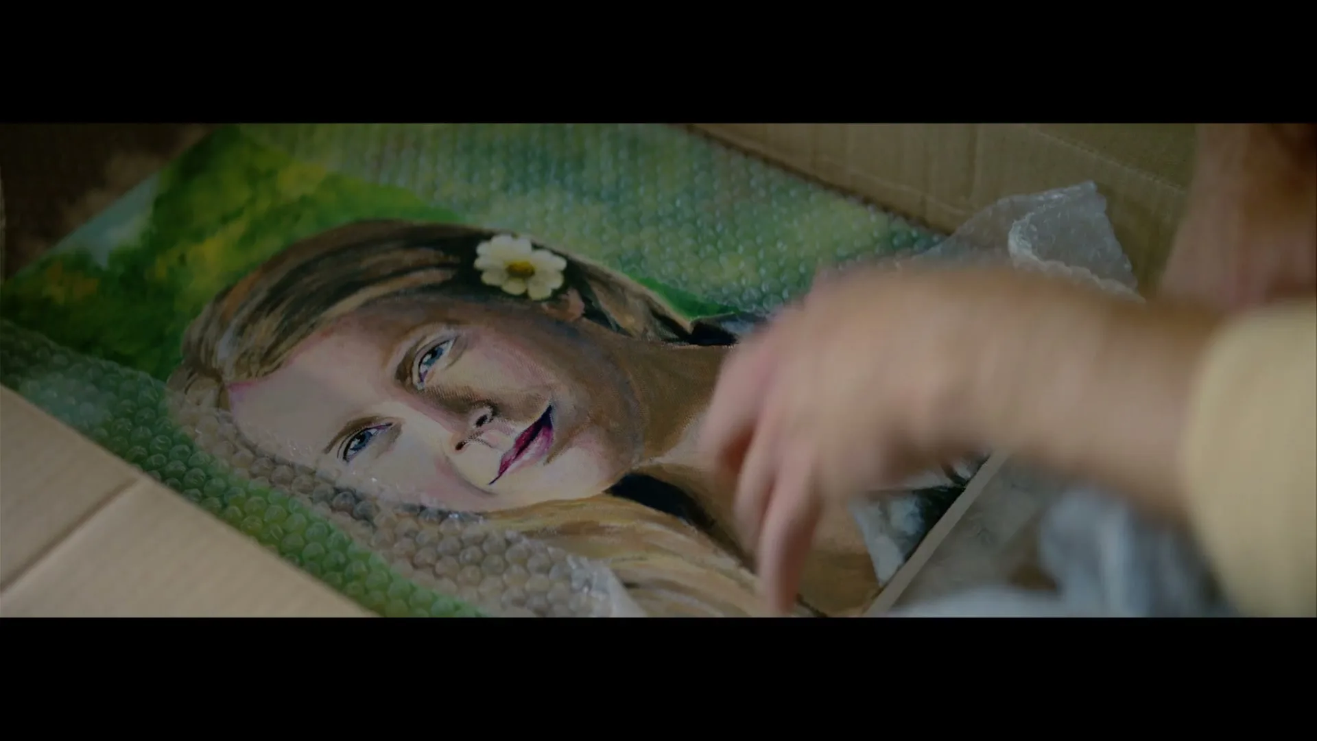A person's hands carefully remove bubble wrap from a painting of a young woman with a flower in her hair, lying in a cardboard box.