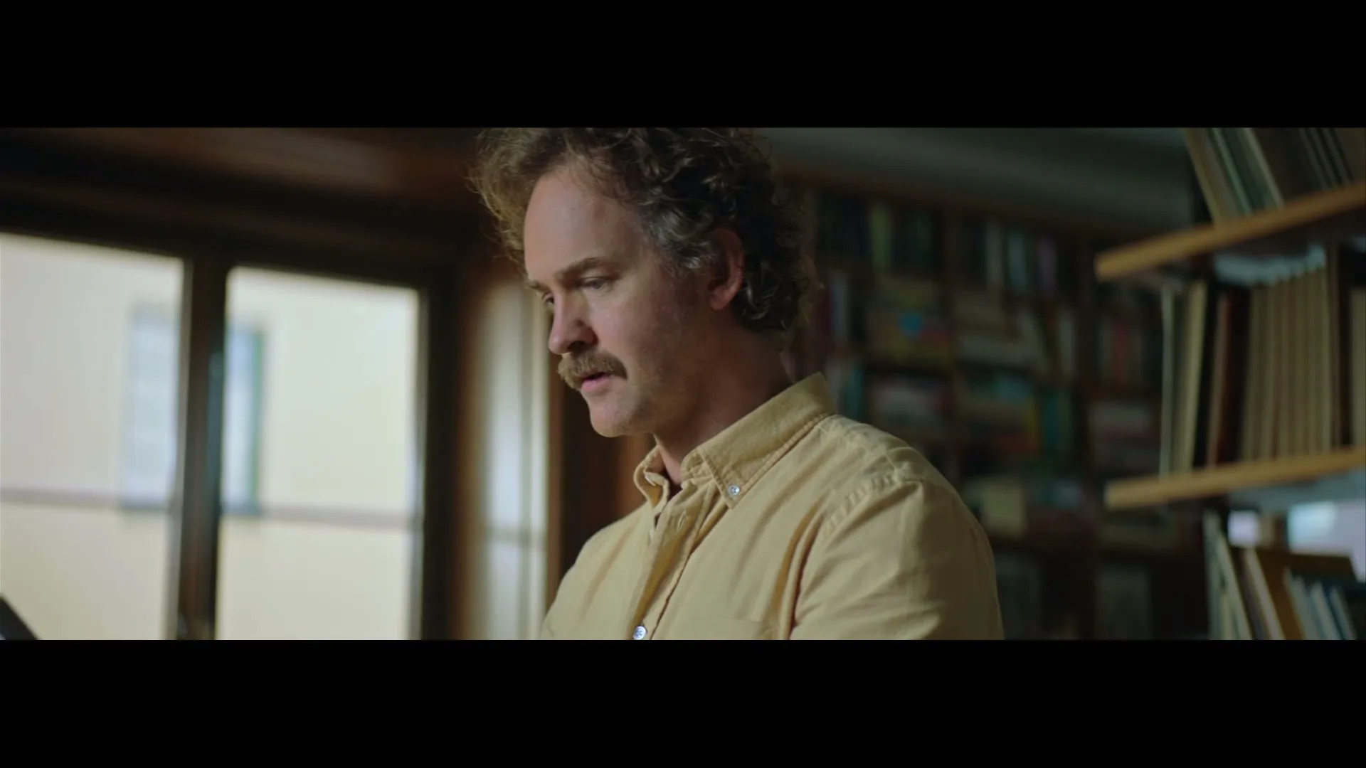 A man with curly hair and a mustache looks down thoughtfully, standing in a room with bookshelves and a window.