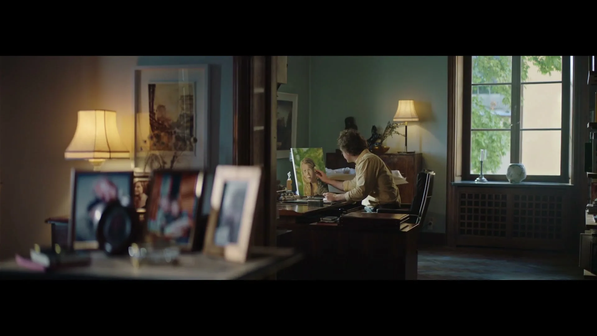 A man with curly hair sits at a desk in a dimly lit room, painting a portrait of a young girl, with framed photos in the foreground.