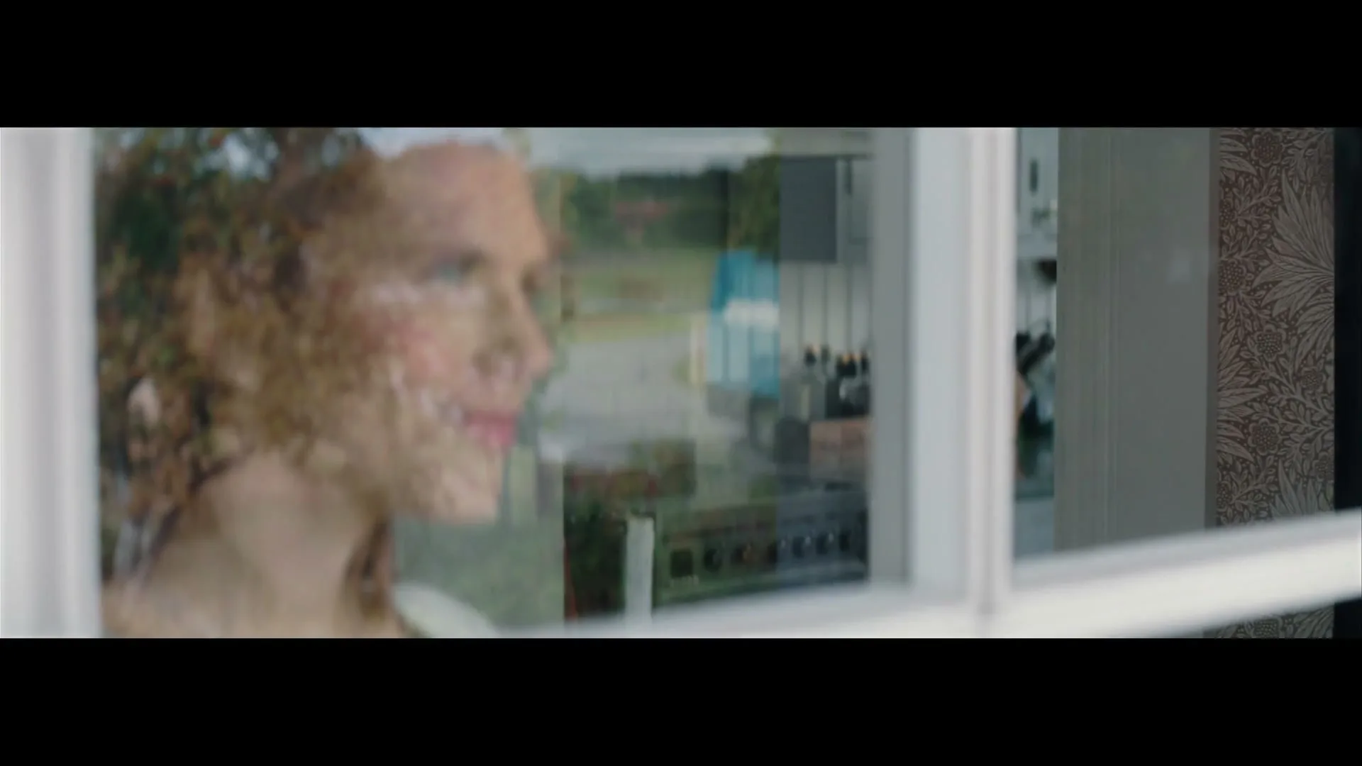 A woman with curly hair looks out a window, reflecting a blue delivery truck driving past a house in a suburban setting.