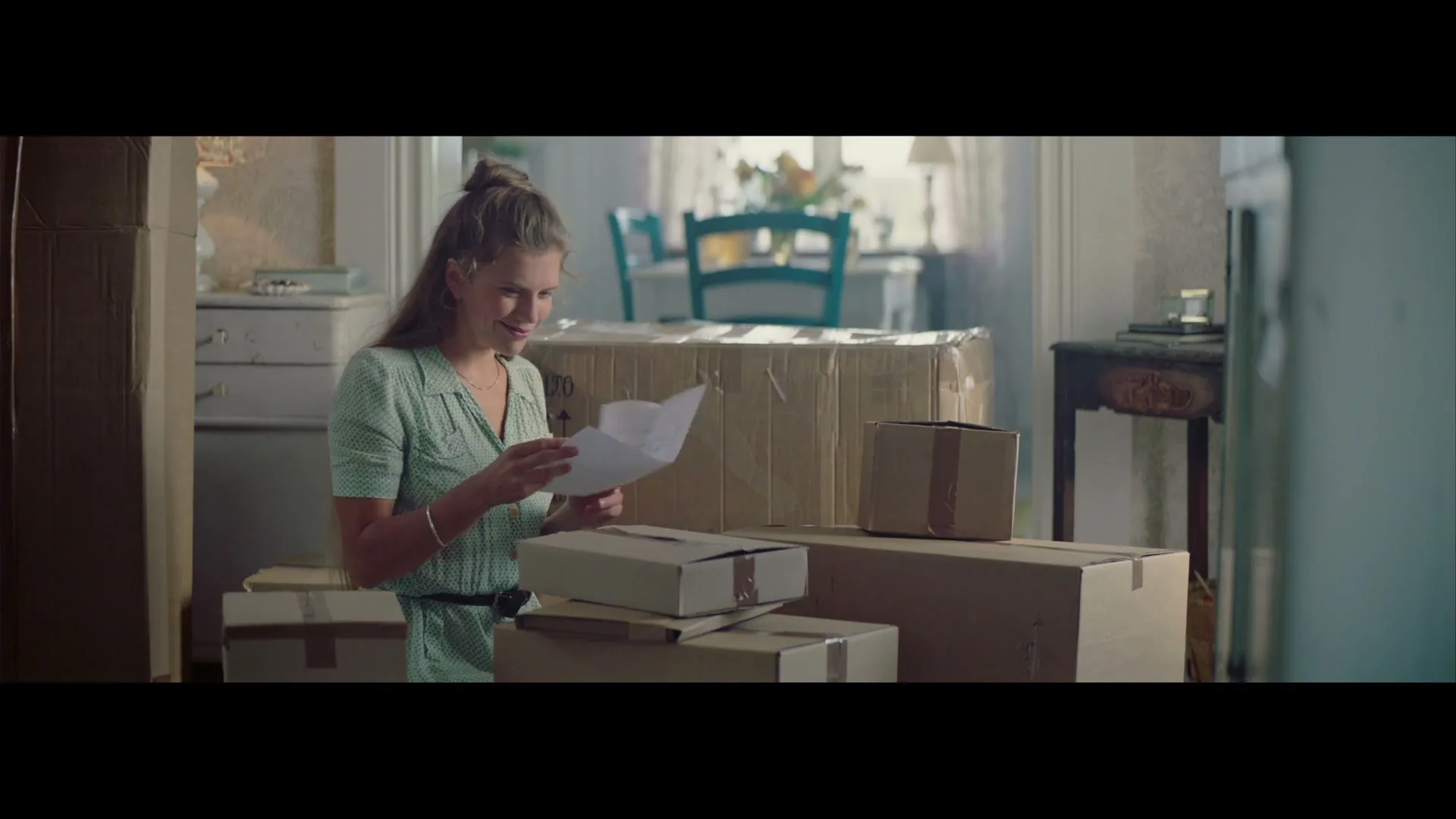 A young woman with her hair in a bun, wearing a green dress, smiles while reading a letter amidst moving boxes in a sunlit room.