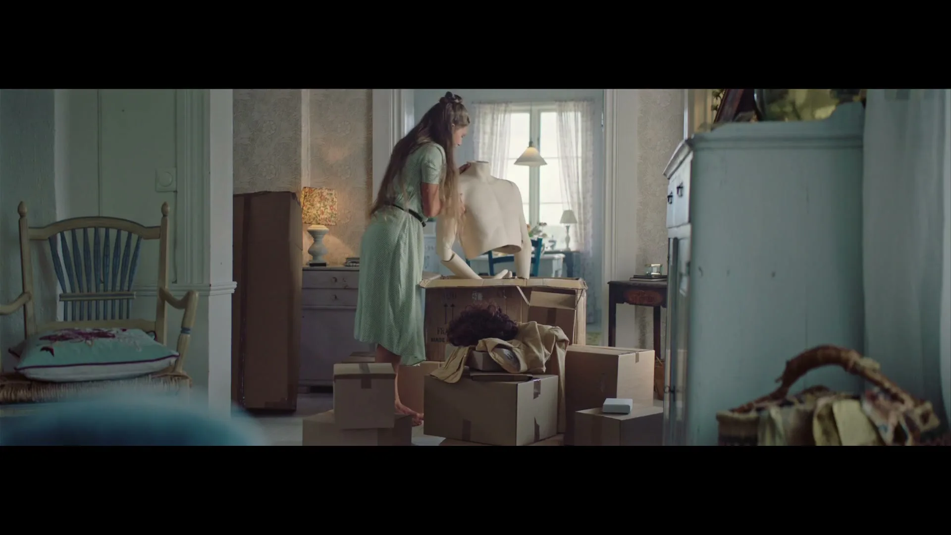 A young woman in a light green dress assembles a mannequin in a sunlit room filled with moving boxes.